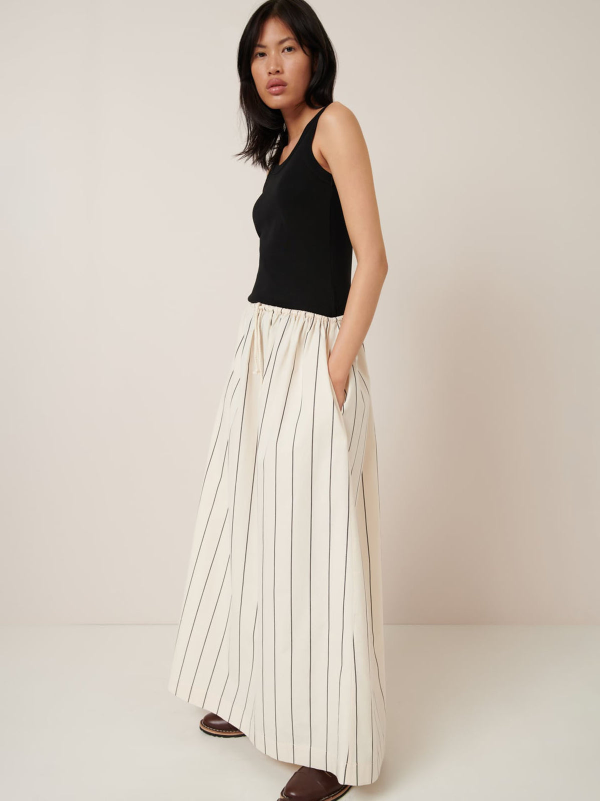 A person models the Kowtow Linear Skirt in Greige Pinstripe, paired with a black sleeveless top and brown shoes against a plain background, highlighting the relaxed fit.