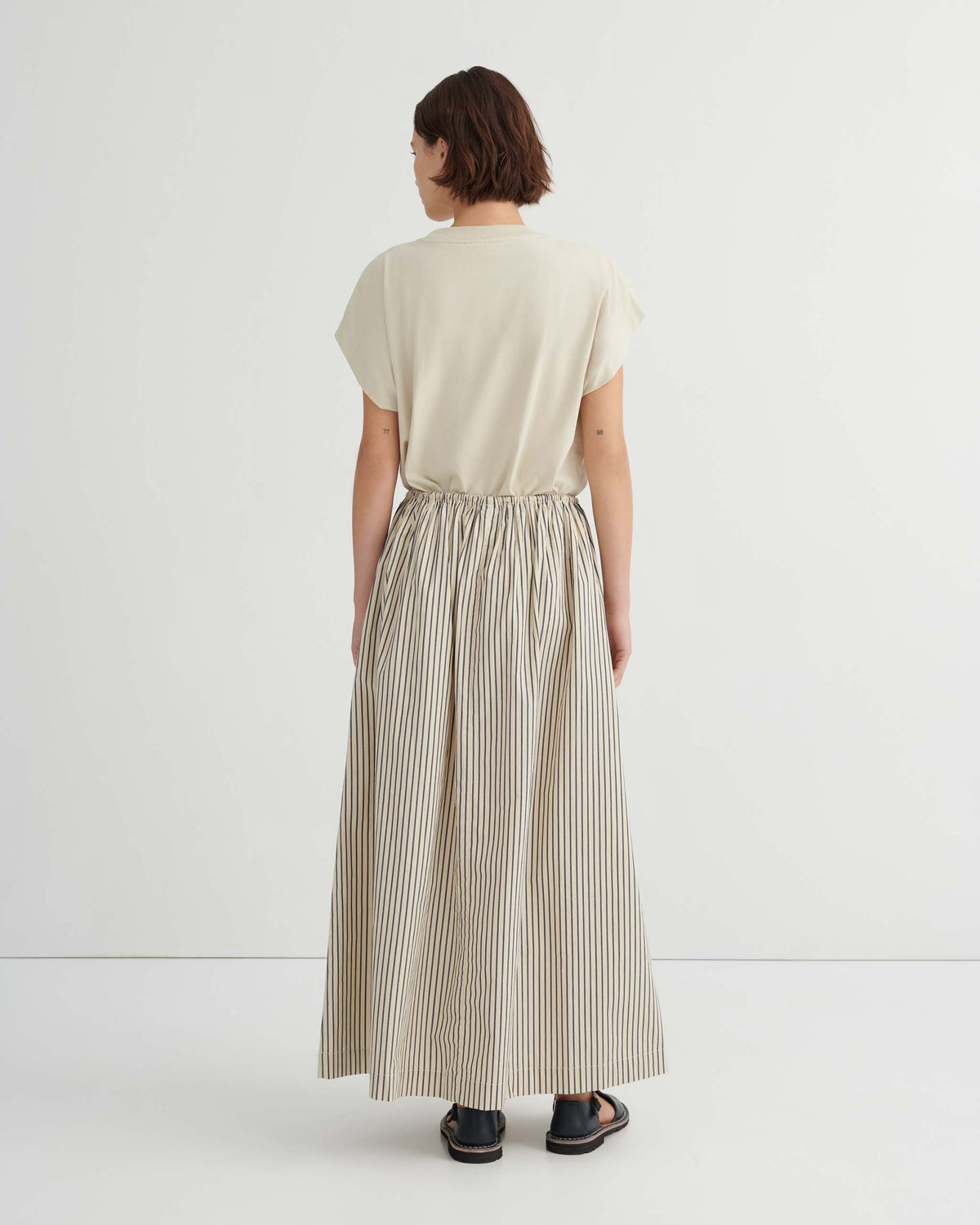 A person seen from behind wearing a beige t-shirt and a long, striped, gathered-waist skirt with black sandals.