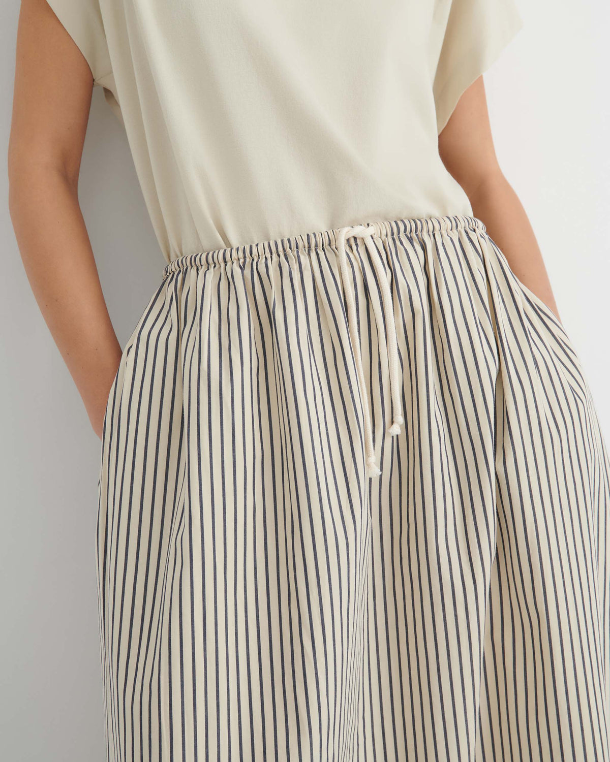 A person wearing a beige short-sleeved top and a white skirt with thin vertical blue stripes and a drawstring waist.