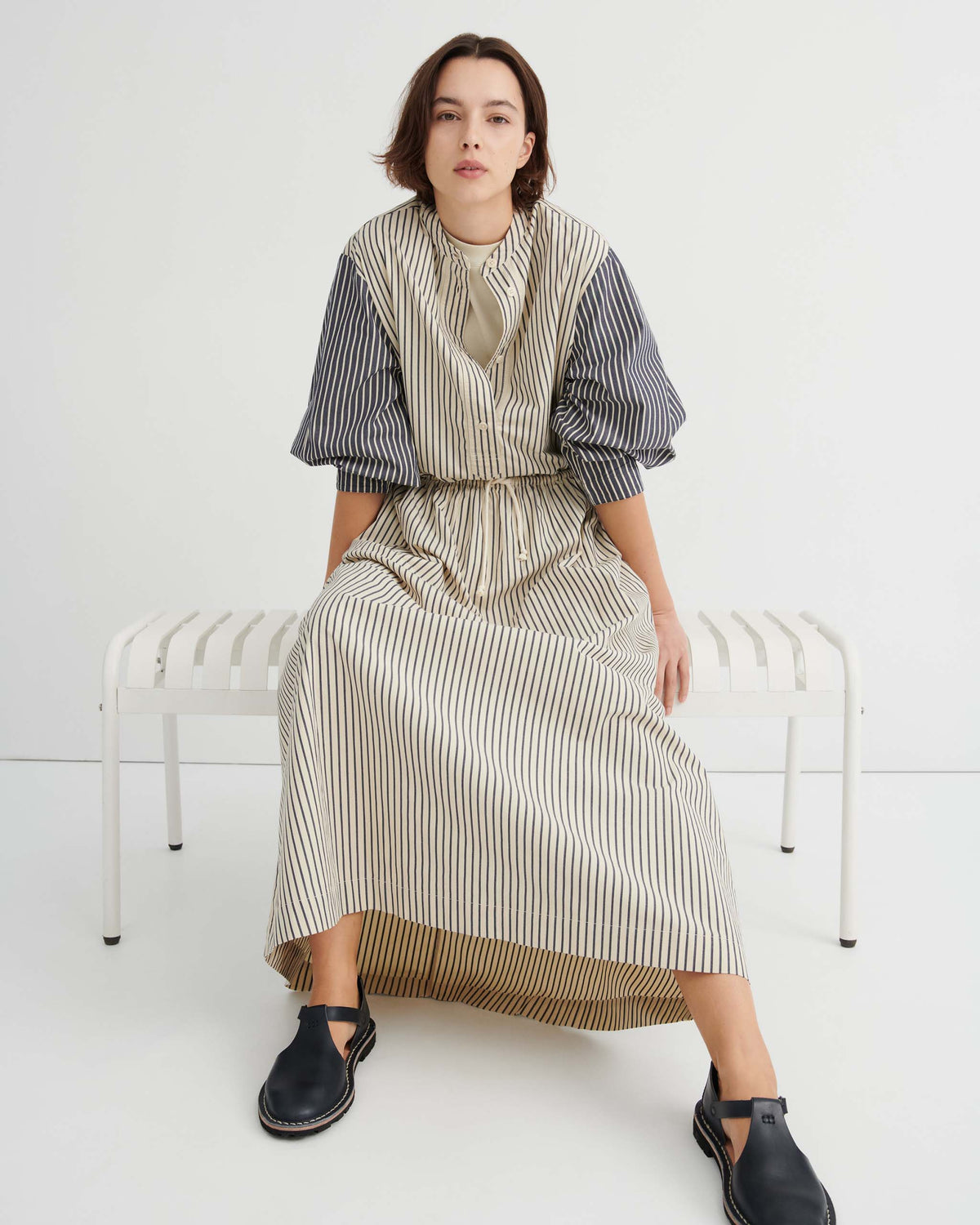 A person sits on a white bench wearing a button-down midi dress with cream and navy stripes and puff sleeves, paired with black leather shoes.