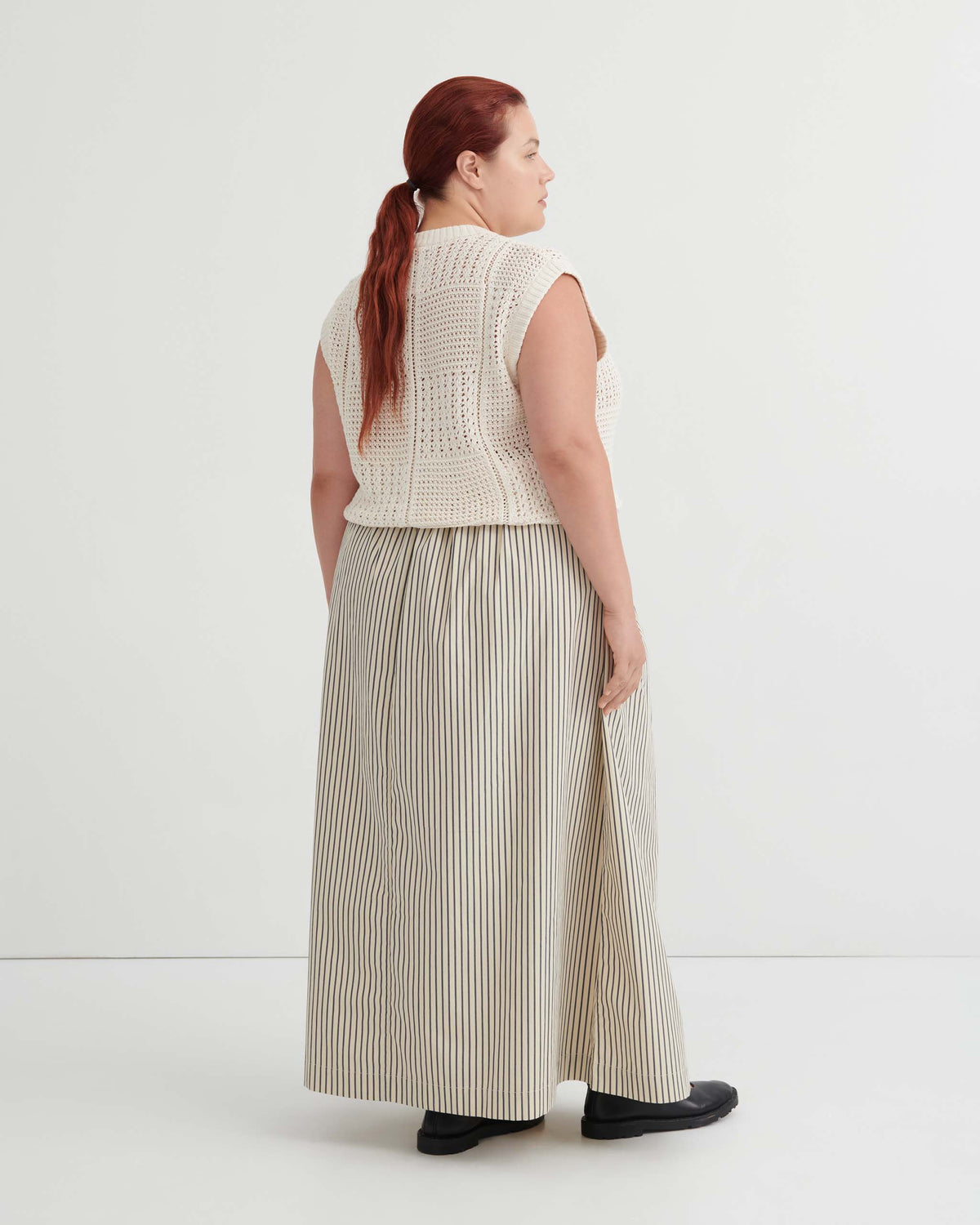 A woman seen from the back wearing a cream knit vest over a long, striped A-line skirt and black shoes.