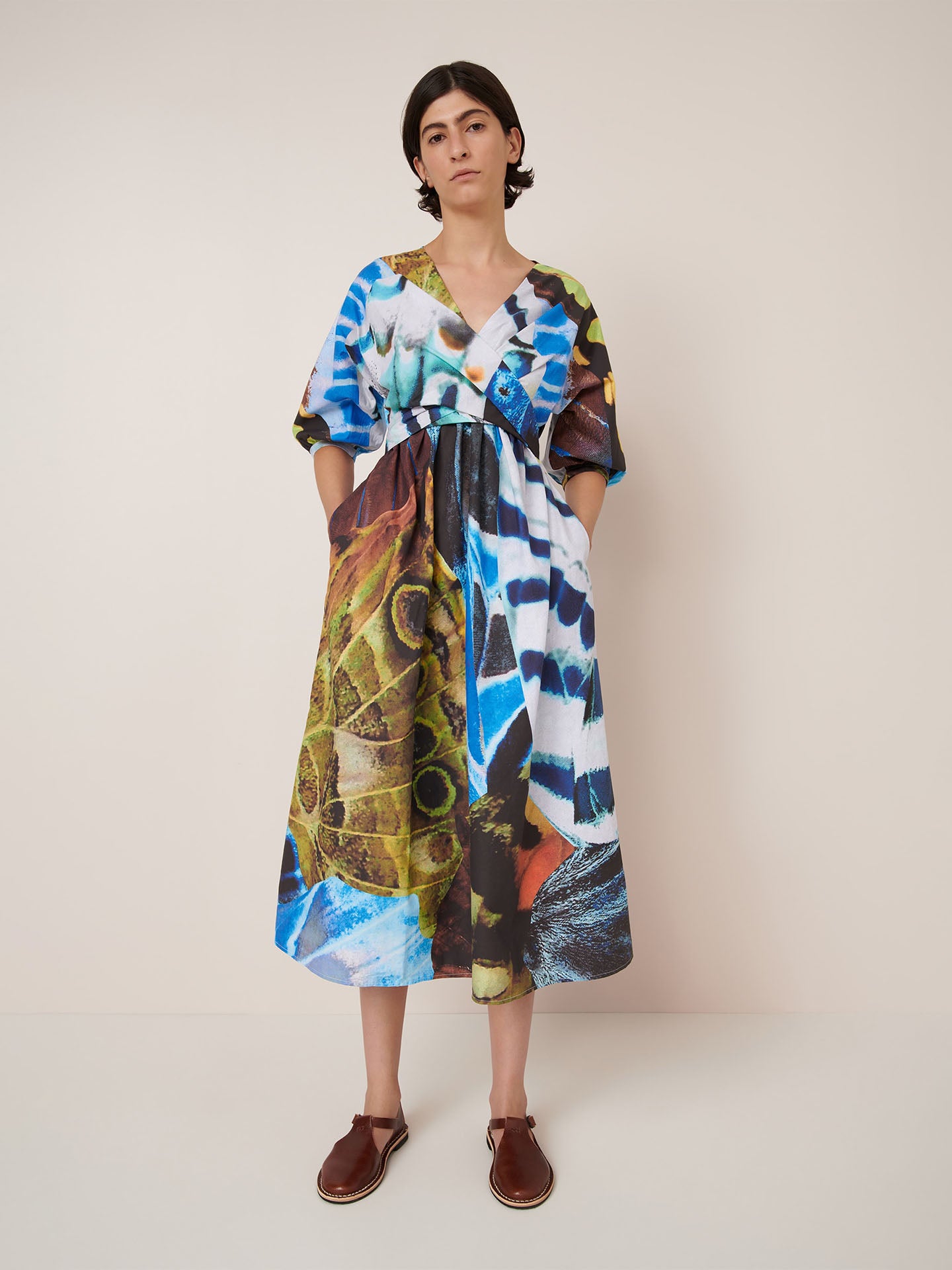 A person stands against a plain wall wearing the Kowtow Marta Dress – Monarch, featuring a bold, multicolored abstract pattern and brown sandals.
