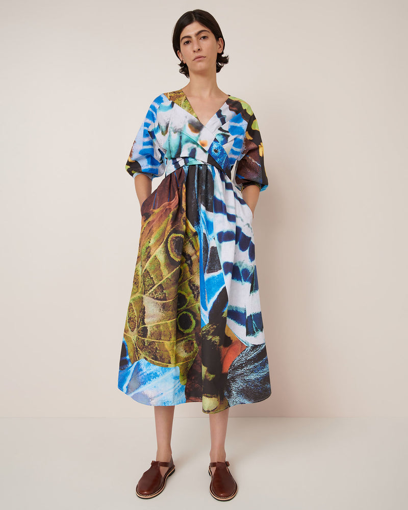 A person stands against a plain wall wearing the Kowtow Marta Dress – Monarch, featuring a bold, multicolored abstract pattern and brown sandals.