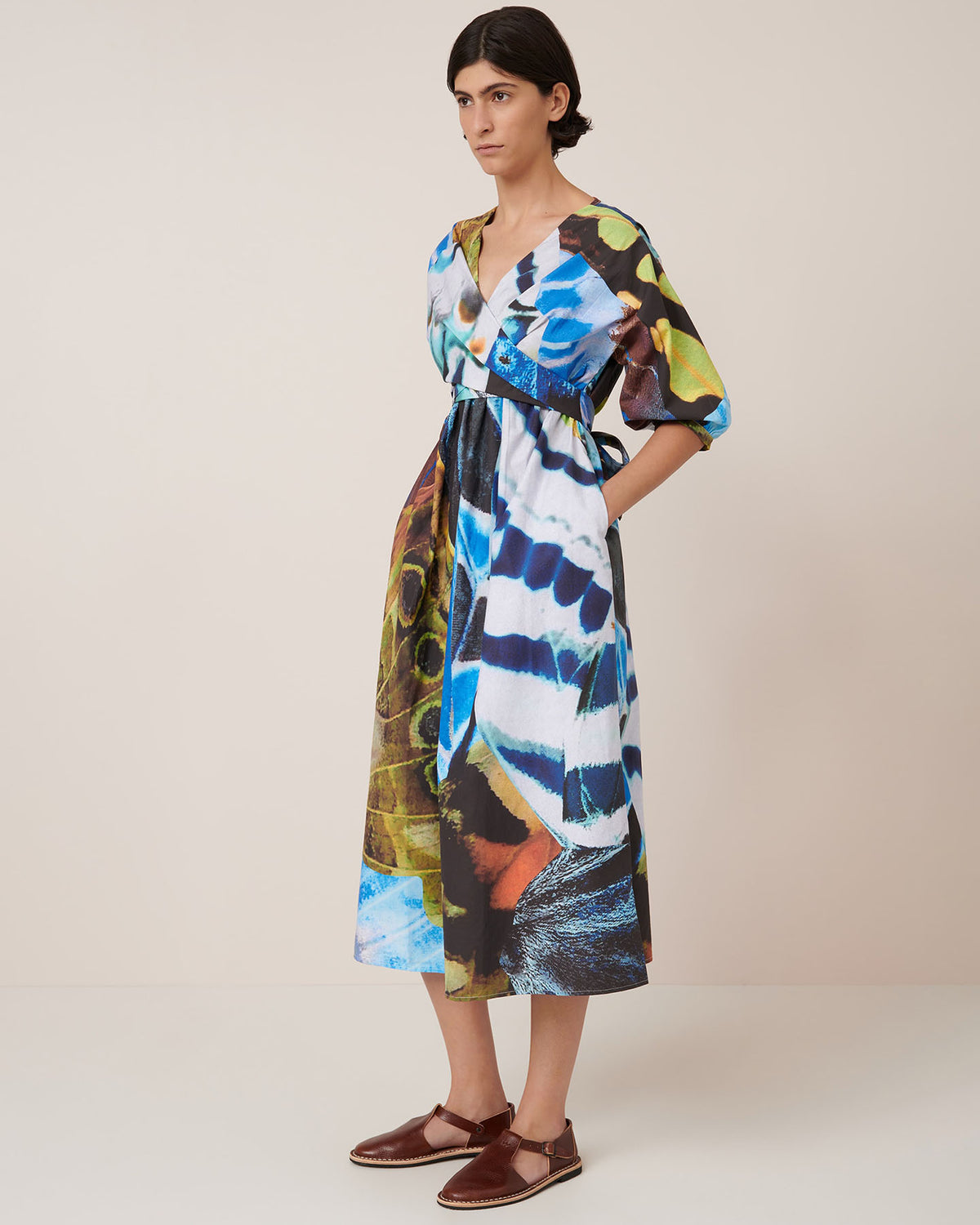 A person stands against a plain background wearing the Kowtow Marta Dress – Monarch, an abstract-patterned adjustable wrap tie dress with half sleeves and a full skirt, paired with brown flat shoes.