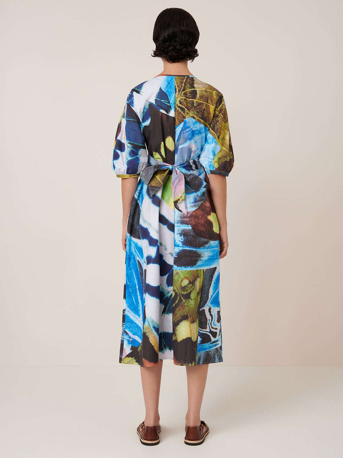 A person faces away, wearing the Kowtow Marta Dress – Monarch, an adjustable wrap tie dress with puff sleeves and a tied back, paired with flat sandals against a plain background.