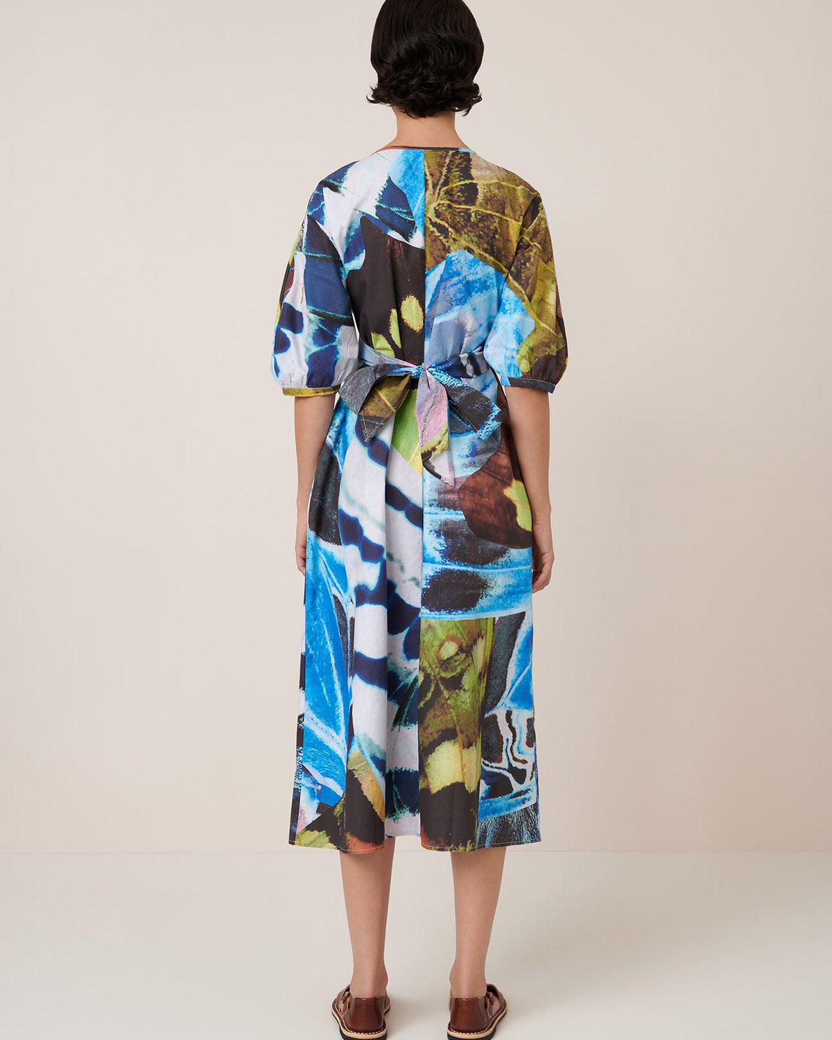 A person faces away, wearing the Kowtow Marta Dress – Monarch, an adjustable wrap tie dress with puff sleeves and a tied back, paired with flat sandals against a plain background.