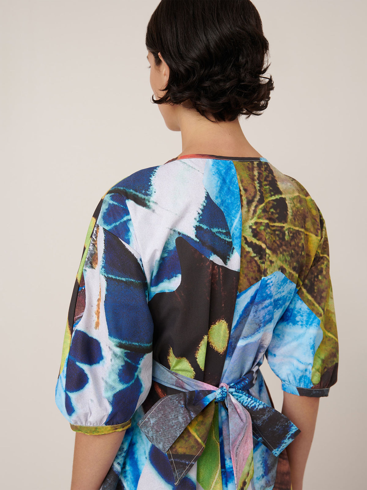 A person with short dark hair is seen from the back wearing the Kowtow Marta Dress – Monarch, an adjustable wrap tie dress with a colorful abstract pattern and a waist bow.