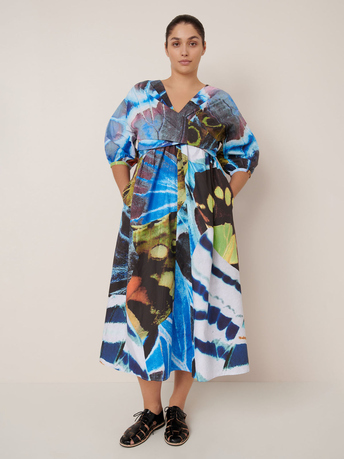 A woman stands against a plain wall wearing the Kowtow Marta Dress – Monarch, featuring a colorful abstract pattern, loose mid-length silhouette, and standard fit, paired with black sandals.