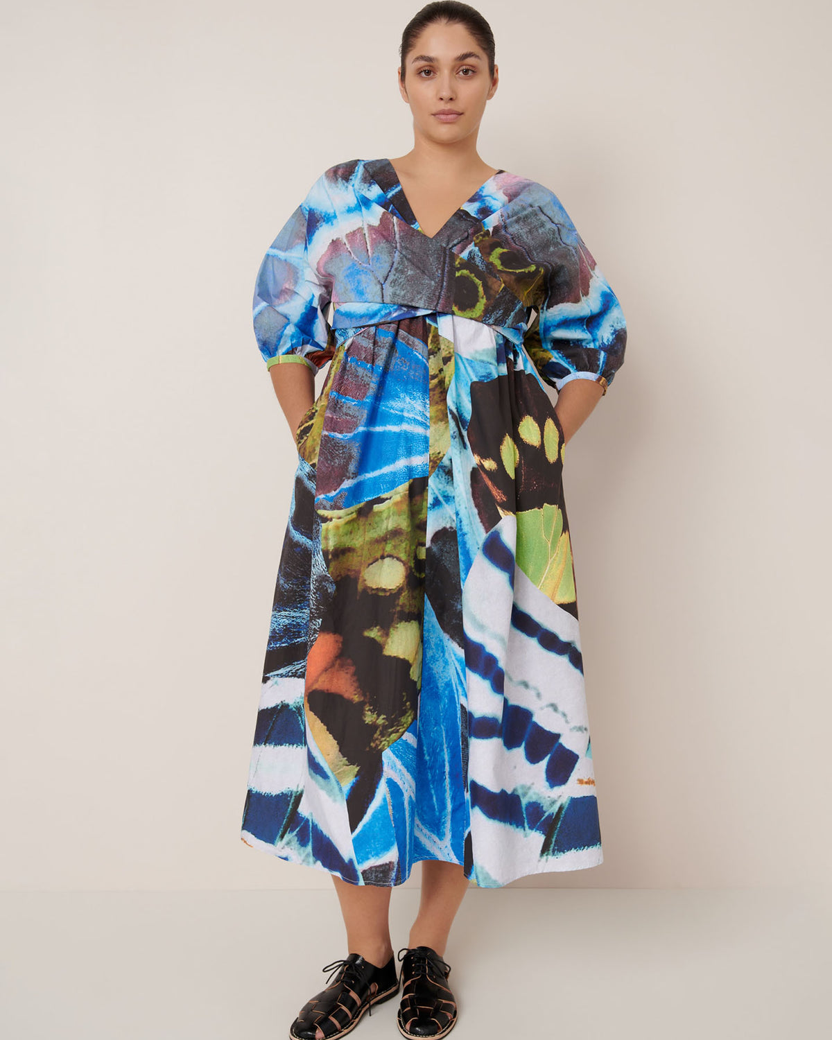 A woman stands against a plain wall wearing the Kowtow Marta Dress – Monarch, featuring a colorful abstract pattern, loose mid-length silhouette, and standard fit, paired with black sandals.