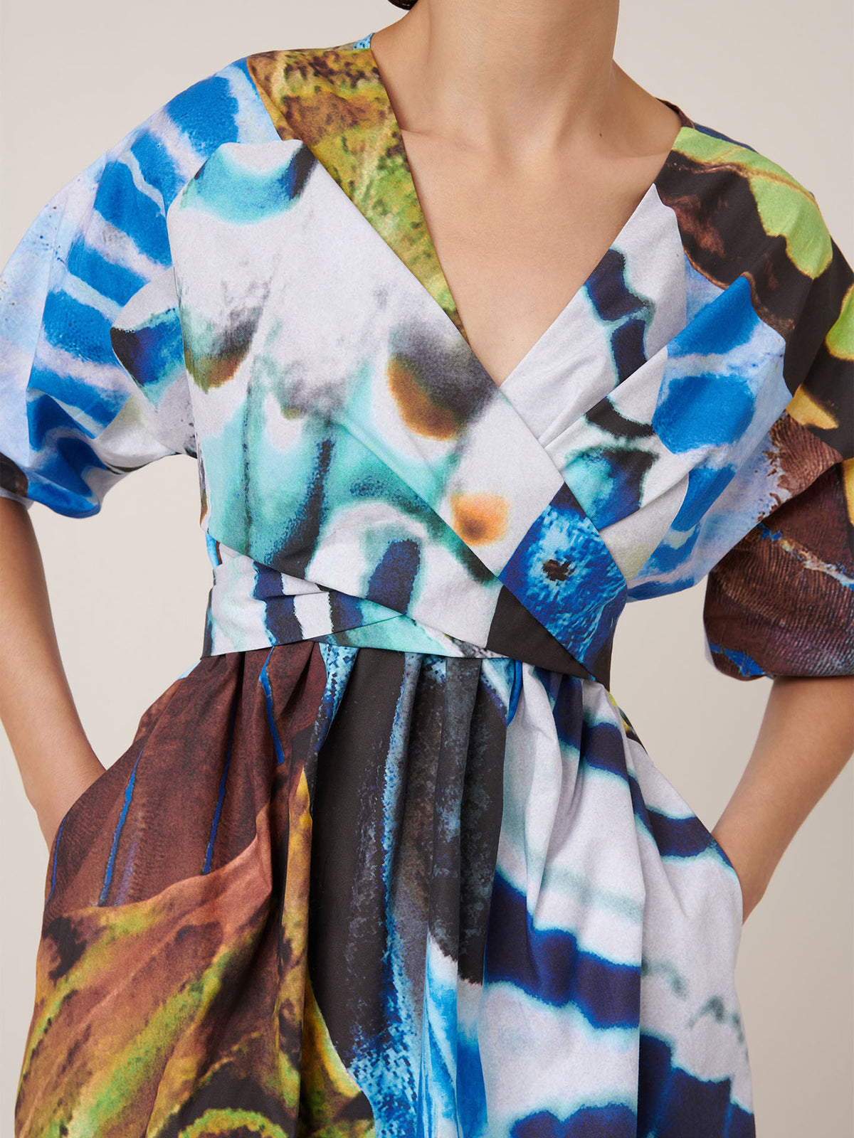 Someone wears the Kowtow Marta Dress – Monarch, a standard fit dress with a V-neck, short sleeves, and a vibrant abstract print in blue, green, brown, black, and white.