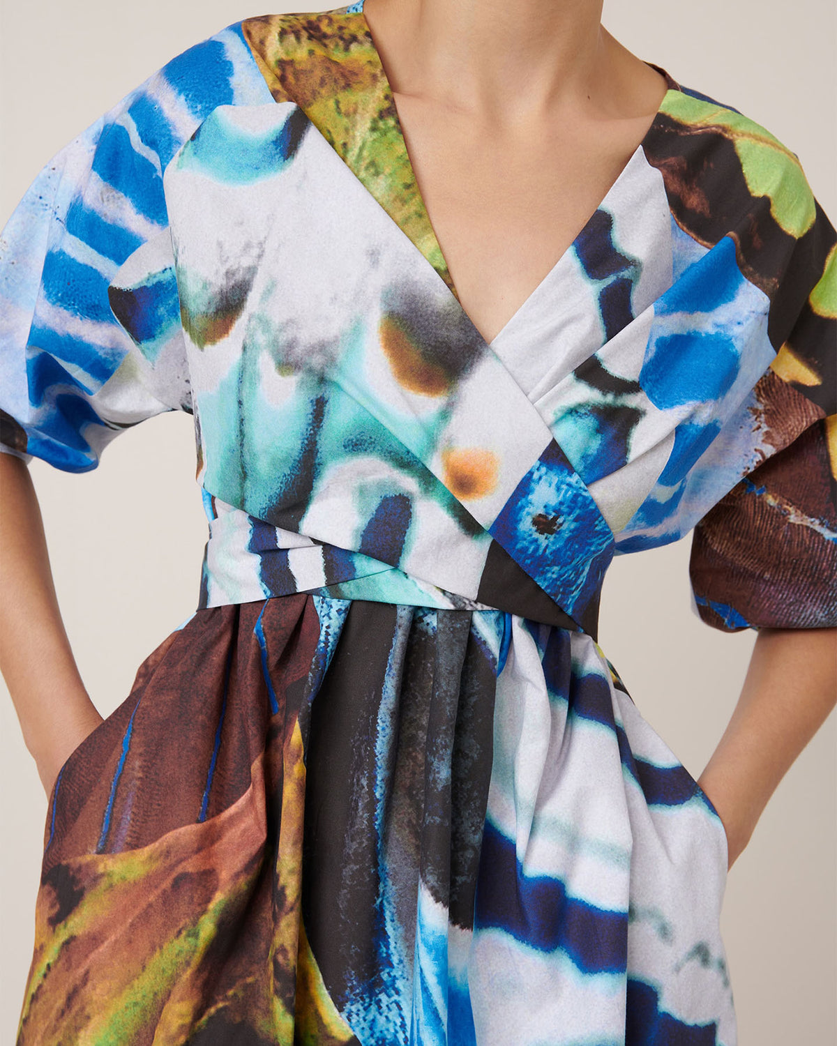 Someone wears the Kowtow Marta Dress – Monarch, a standard fit dress with a V-neck, short sleeves, and a vibrant abstract print in blue, green, brown, black, and white.