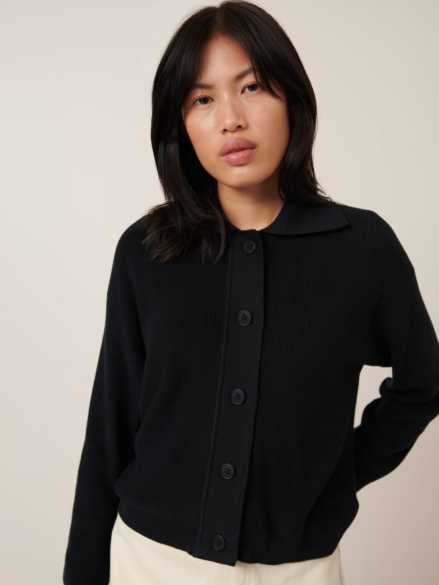 Someone is wearing the Merel Cardigan in black by Kowtow, styled as a button-up with a standard fit, paired with white pants against a plain backdrop.