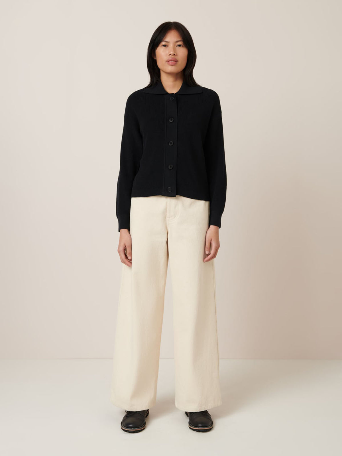 A person stands against a plain background wearing Kowtow&#39;s Merel Cardigan in black, paired with cream wide-leg pants and black shoes, effortlessly showcasing a standard fit with style.