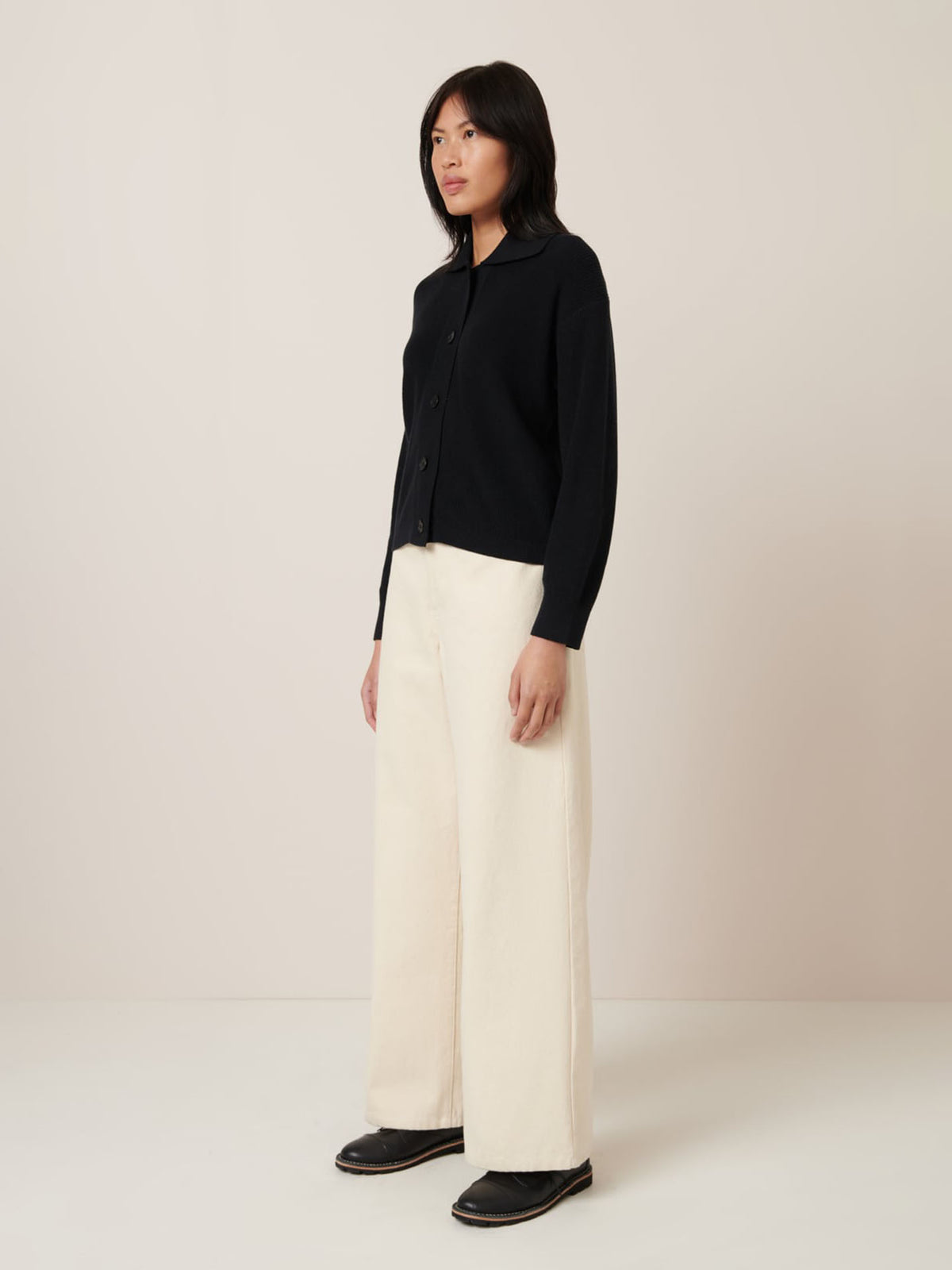 A person wearing a Kowtow Merel Cardigan in black paired with wide-legged white pants stands against a neutral backdrop, presenting a chic silhouette. Consult the size guide for precise measurements to ensure your perfect fit.