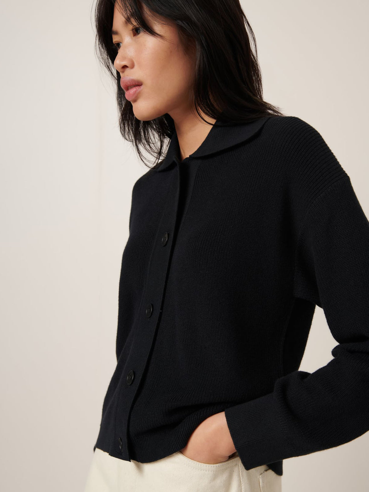 A person with long dark hair models the Kowtow Merel Cardigan in black, featuring a standard fit. They wear light-colored pants and stand sideways with hands in pockets against a plain background, embodying design that fits any size guide effortlessly.