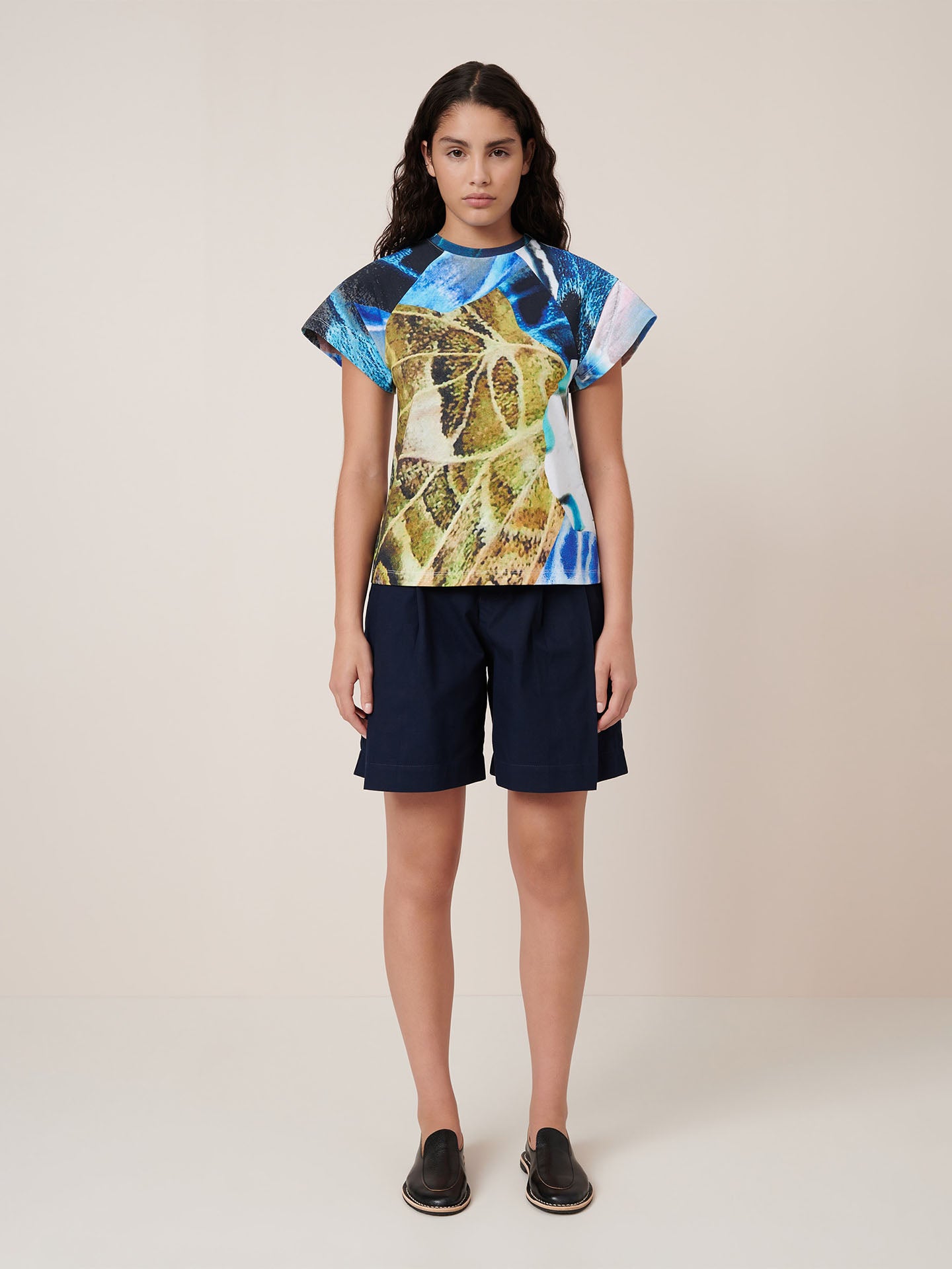 A person stands facing forward in the Kowtow Monarch Top, a colorful abstract-patterned standard fit t-shirt, paired with dark knee-length shorts and black slip-on shoes against a plain background.