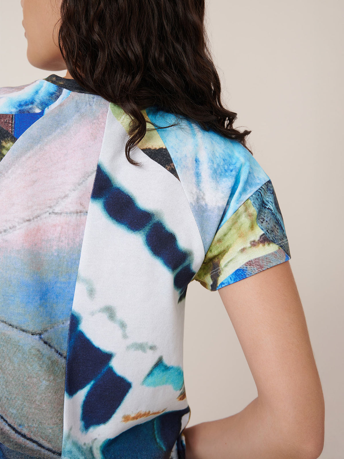 A person with dark, wavy hair wears the Kowtow Monarch Top, a short-sleeve shirt with a colorful abstract pattern in blue, green, and white. The image highlights the standard fit on the back and shoulder of the shirt.