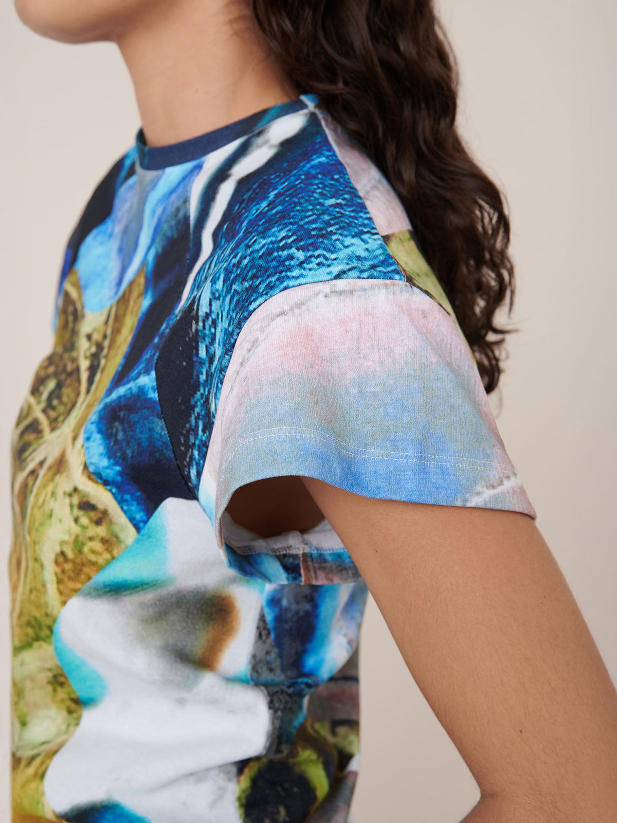 A person with long, dark, wavy hair is shown from the side wearing the Kowtow Monarch Top, a standard-fit short-sleeved shirt featuring a multicolored abstract print.