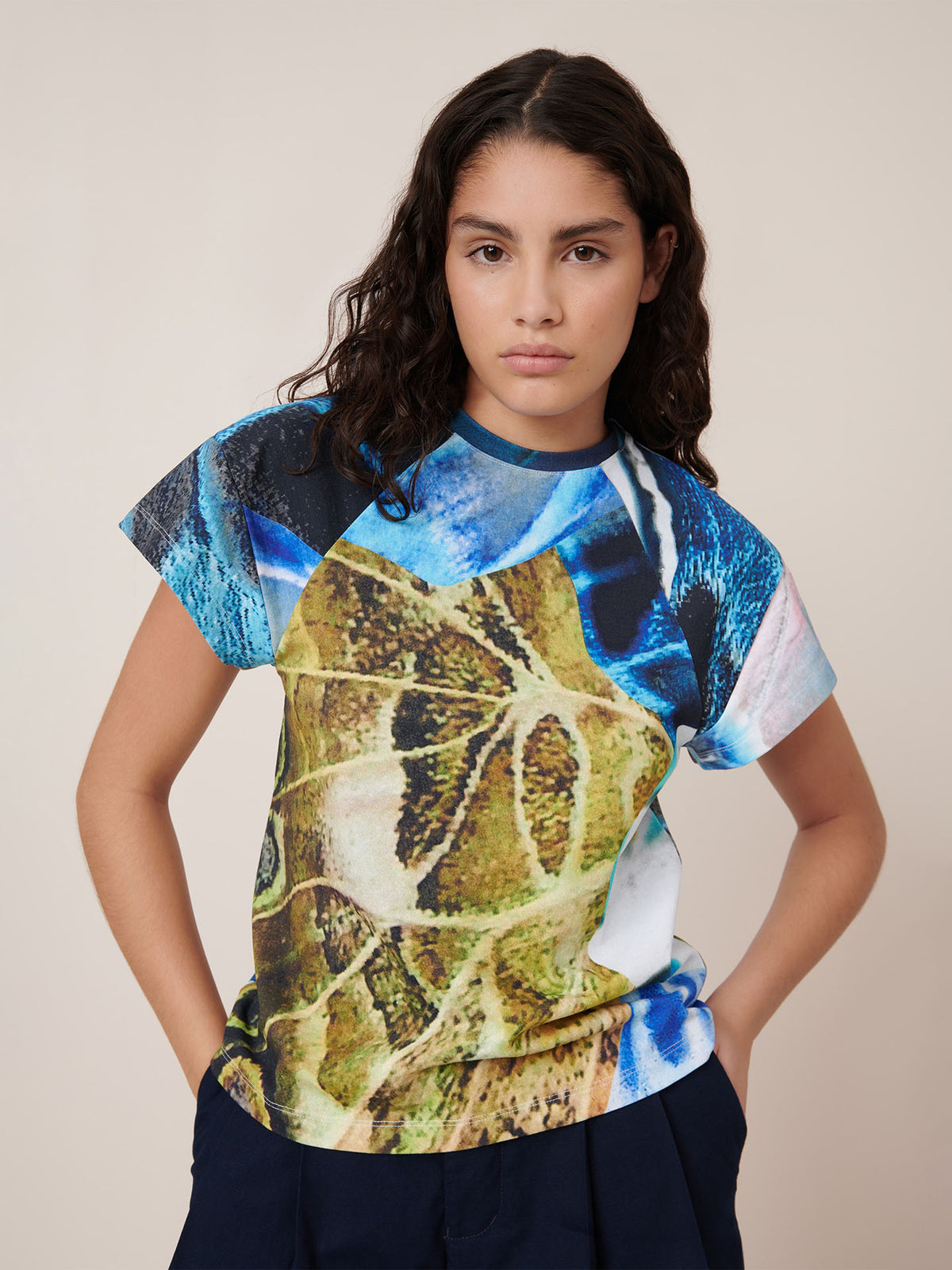 A person with long dark hair wears the Kowtow Monarch Top, a colorful graphic tee with abstract blue and yellow patterns. It has a standard fit that’s true to size and is pictured against a plain light background with hands in pockets.