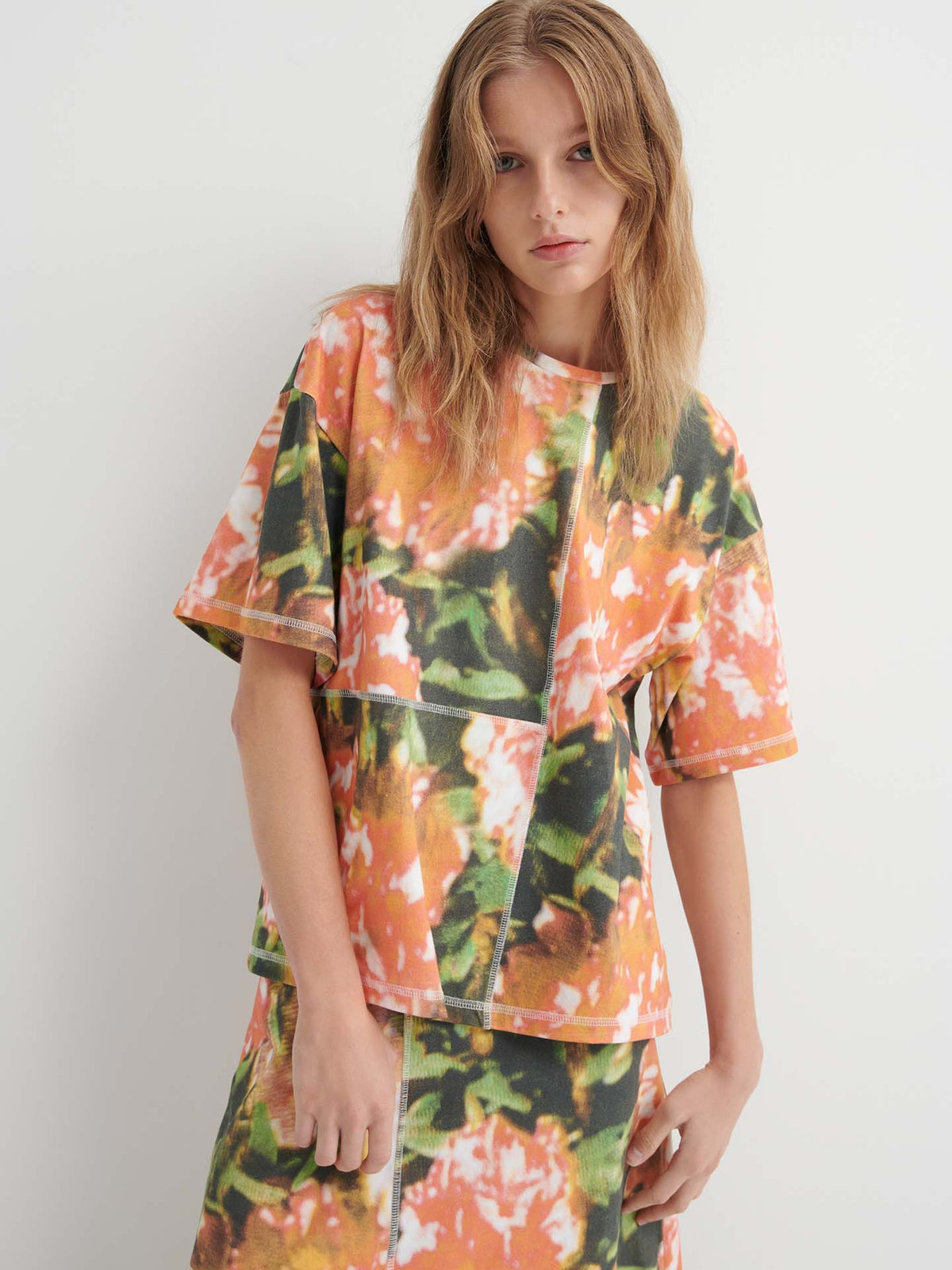 A person wears the Montage Tee – Flower Book, a relaxed fit short-sleeved top with an abstract orange, green, and white floral print, paired with a matching skirt, standing against a plain white background.