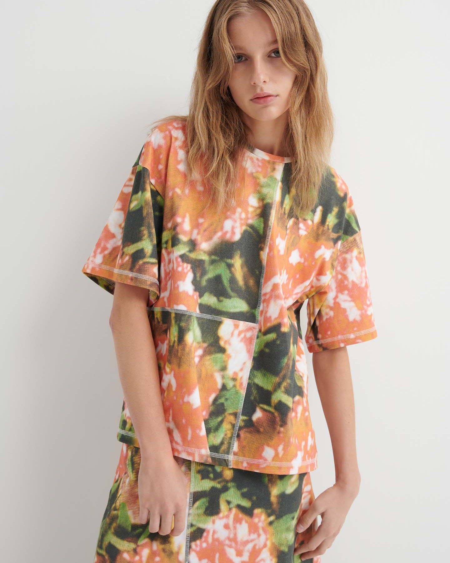 A person wears the Montage Tee – Flower Book, a relaxed fit short-sleeved top with an abstract orange, green, and white floral print, paired with a matching skirt, standing against a plain white background.