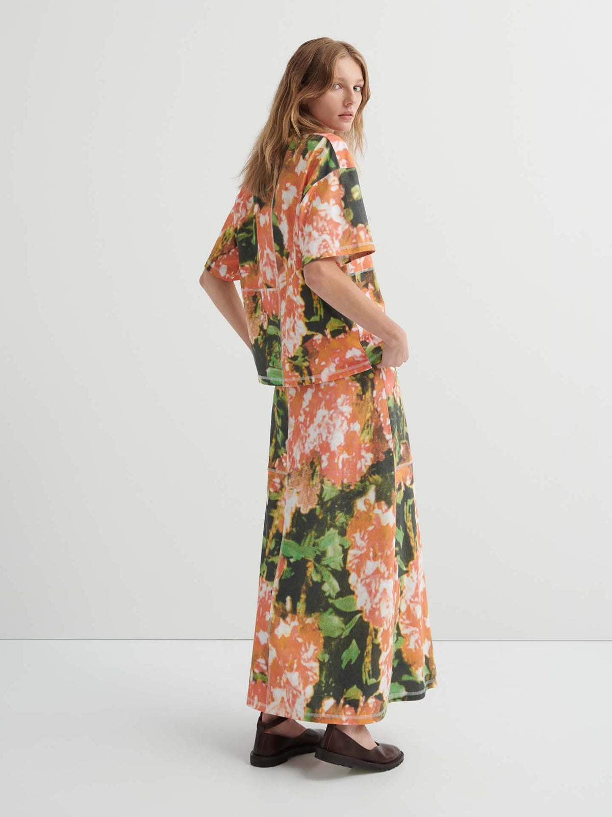 A woman stands in profile wearing the Montage Tee – Flower Book, a relaxed fit floral-patterned top, paired with a matching long skirt and brown shoes, set against a plain white background.