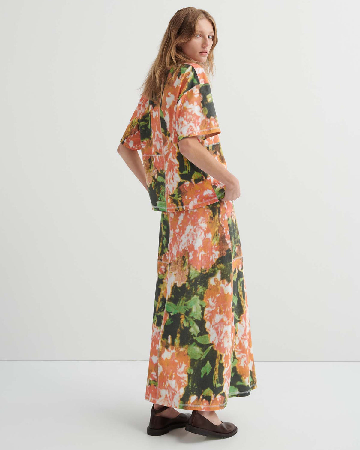 A woman stands in profile wearing the Montage Tee – Flower Book, a relaxed fit floral-patterned top, paired with a matching long skirt and brown shoes, set against a plain white background.