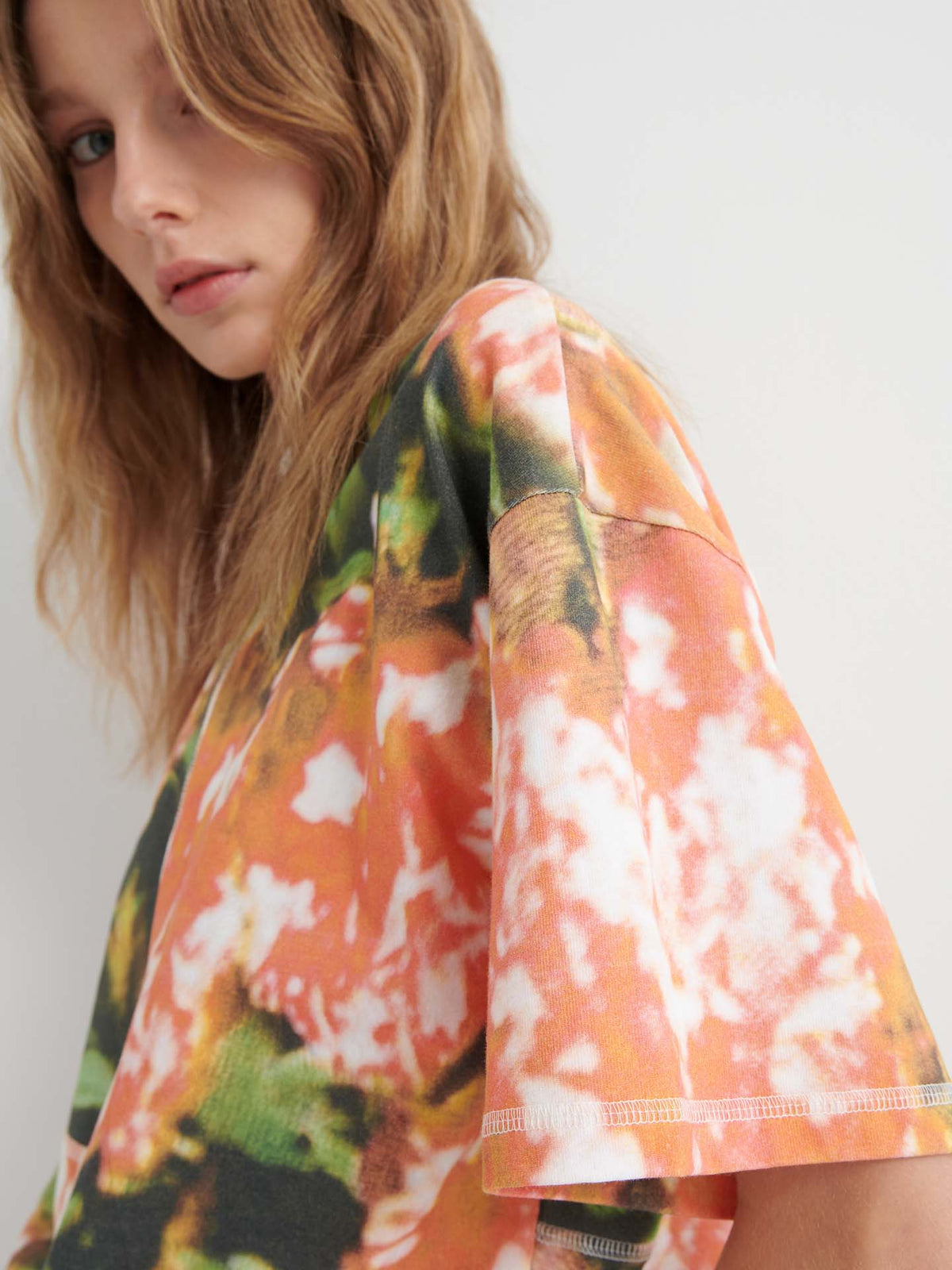 A person with wavy light brown hair wears the Montage Tee – Flower Book in a relaxed fit, featuring short sleeves and an abstract orange, green, and white pattern that stands out against a plain background.