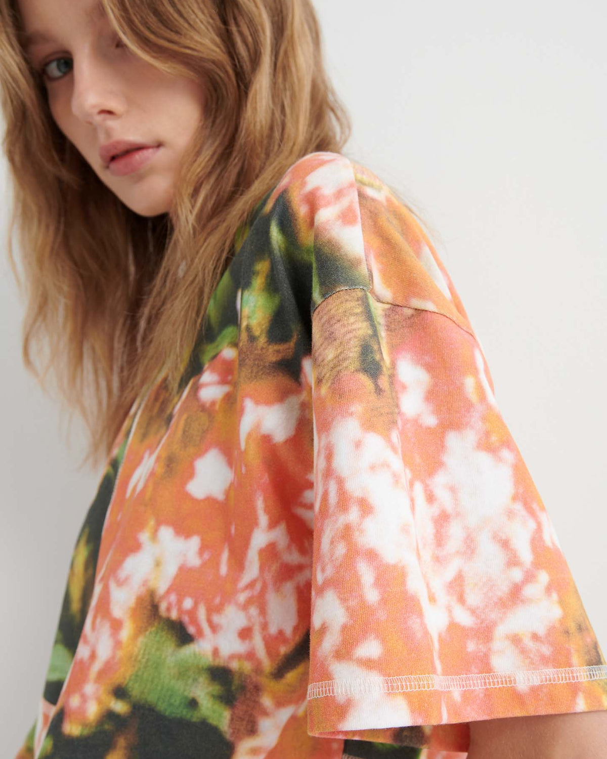 A person with wavy light brown hair wears the Montage Tee – Flower Book in a relaxed fit, featuring short sleeves and an abstract orange, green, and white pattern that stands out against a plain background.