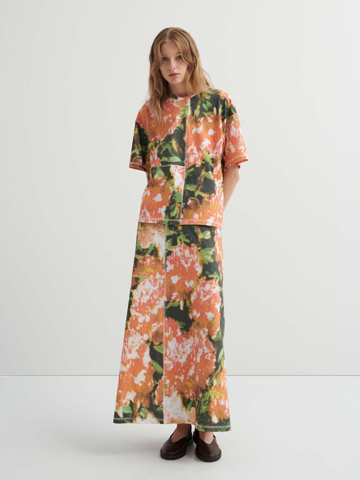 A person stands against a plain background wearing a relaxed fit Montage Tee – Flower Book and a long skirt with an abstract orange floral pattern, paired with dark flat shoes.