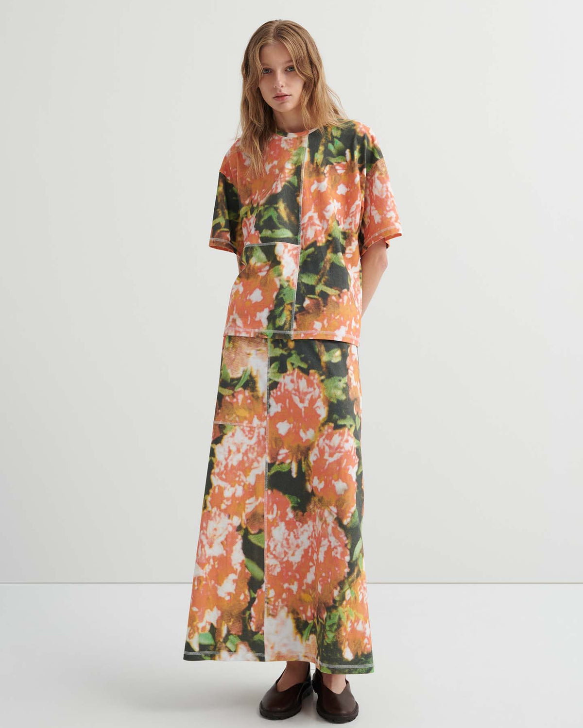 A person stands against a plain background wearing a relaxed fit Montage Tee – Flower Book and a long skirt with an abstract orange floral pattern, paired with dark flat shoes.