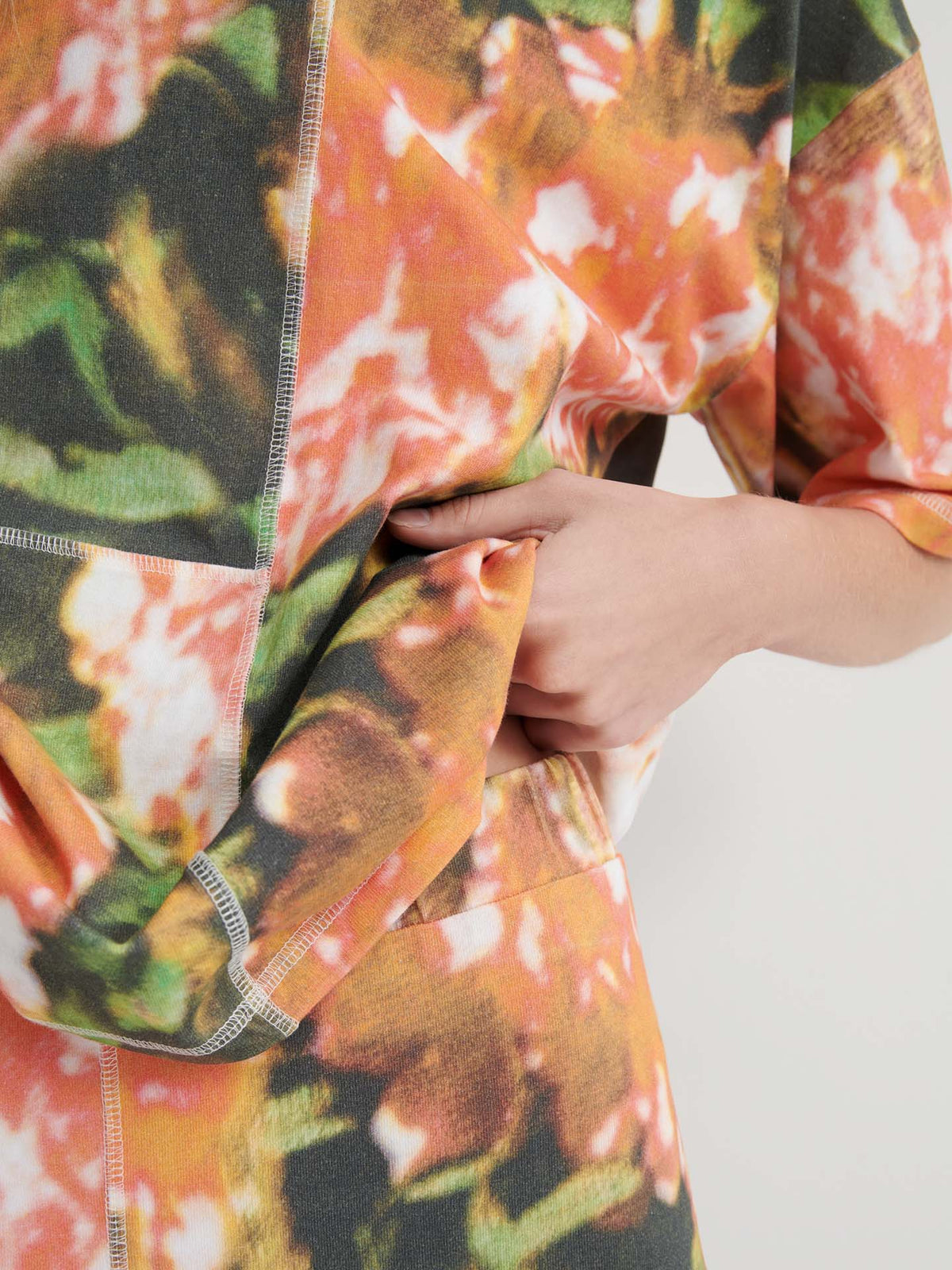 A person wearing the Montage Tee – Flower Book, featuring a colorful abstract pattern and relaxed fit, holds part of their shirt with one hand, partially revealing the side of their torso.