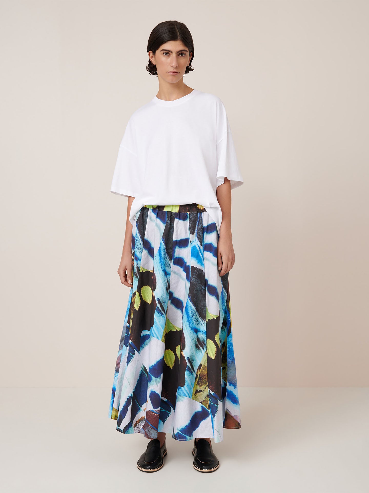 A person stands against a plain background wearing a relaxed fit white t-shirt, black shoes, and the Kowtow Mosaic Skirt – Monarch, a long high-waist skirt with a colorful abstract pattern.