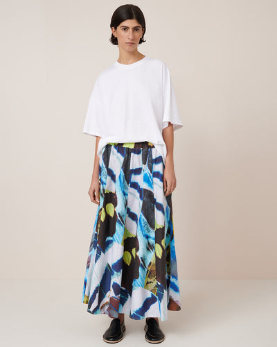 A person stands against a plain background wearing a relaxed fit white t-shirt, black shoes, and the Kowtow Mosaic Skirt – Monarch, a long high-waist skirt with a colorful abstract pattern.