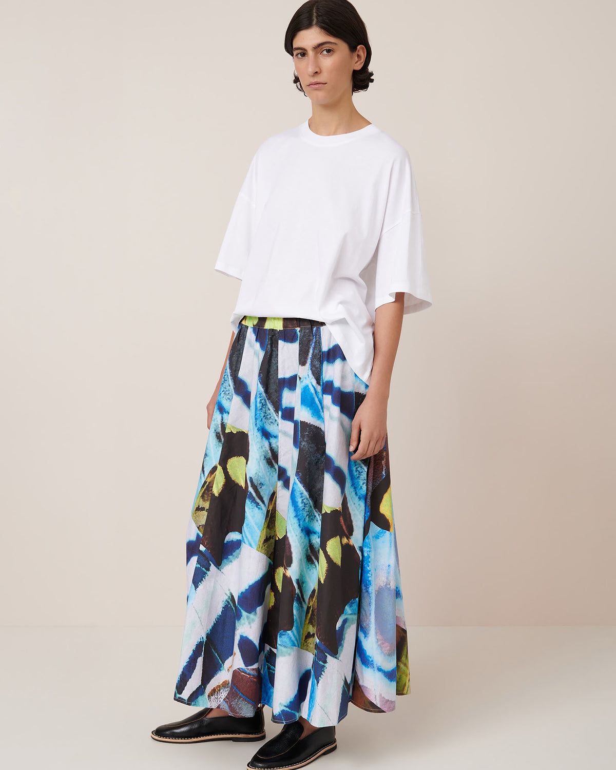 Person wears Kowtow’s Mosaic Skirt – Monarch, a high-waist, wide skirt with bold abstract blue, black, and yellow patterns, paired with a loose white t-shirt against a beige background. Ideal for those who love a relaxed fit.