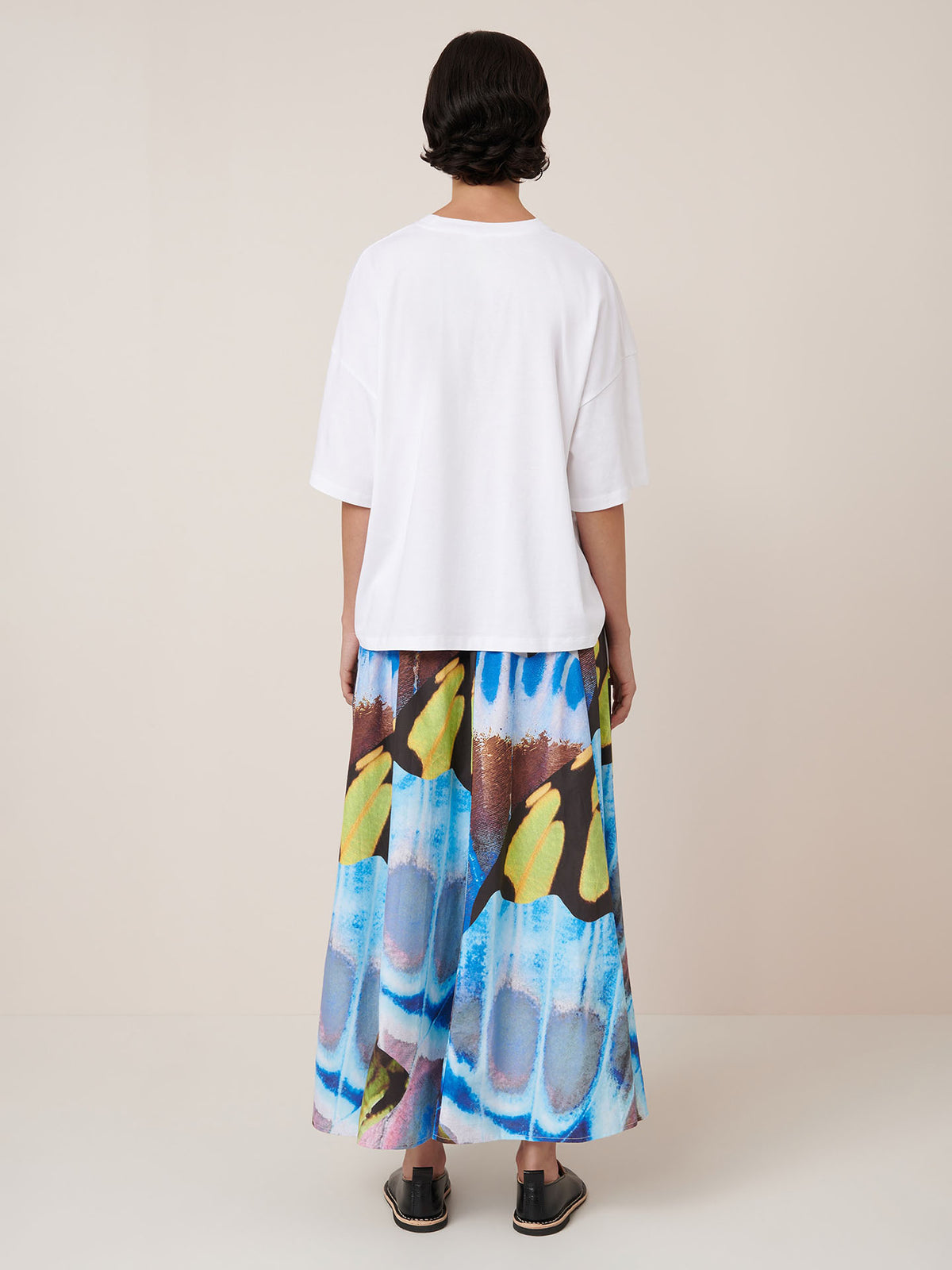 A person wears the Kowtow Mosaic Skirt – Monarch, a high-waisted, colorful patterned maxi skirt, with a plain white tee and black sandals against a neutral backdrop. Check the size guide for your ideal relaxed fit.