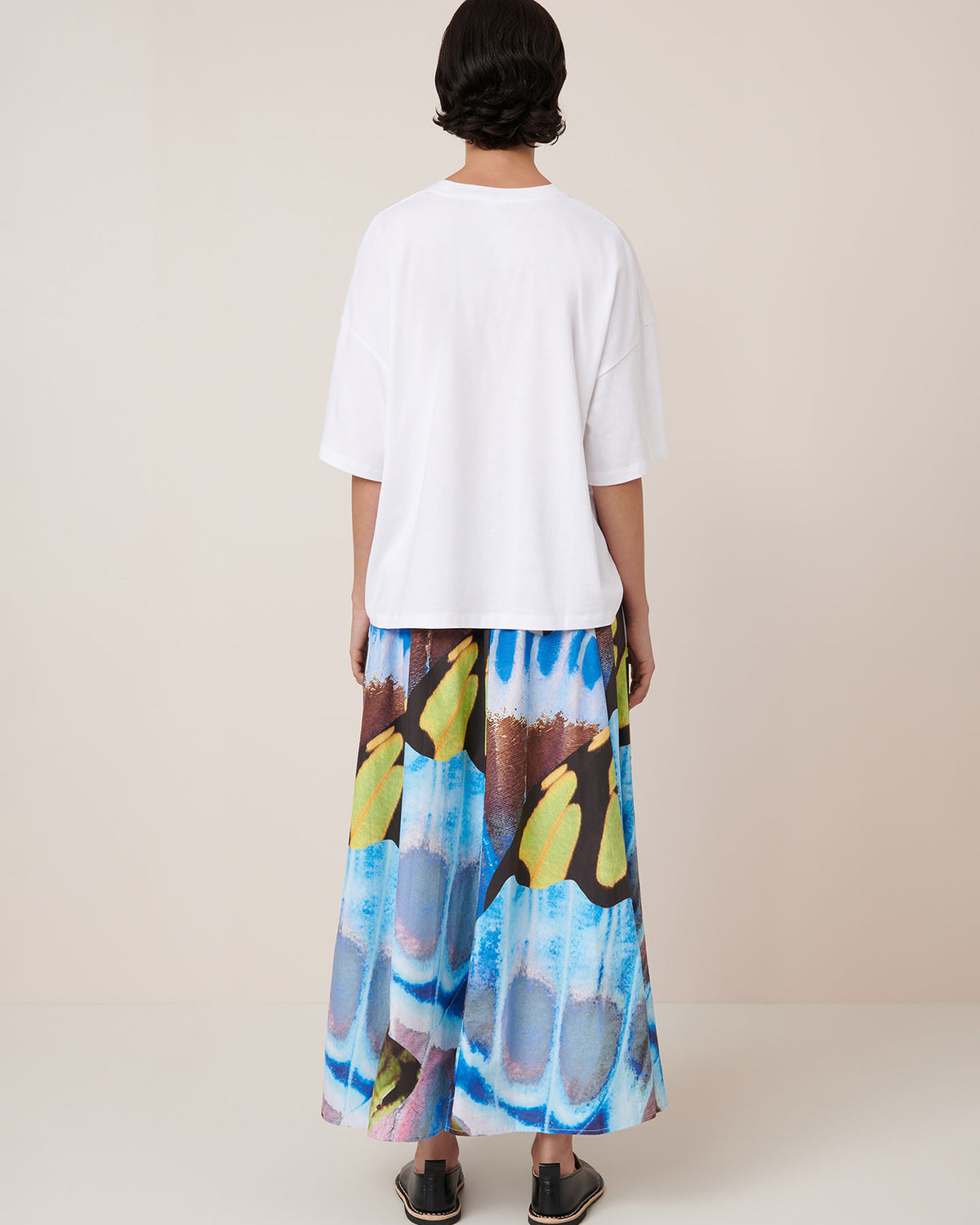 A person wears the Kowtow Mosaic Skirt – Monarch, a high-waisted, colorful patterned maxi skirt, with a plain white tee and black sandals against a neutral backdrop. Check the size guide for your ideal relaxed fit.