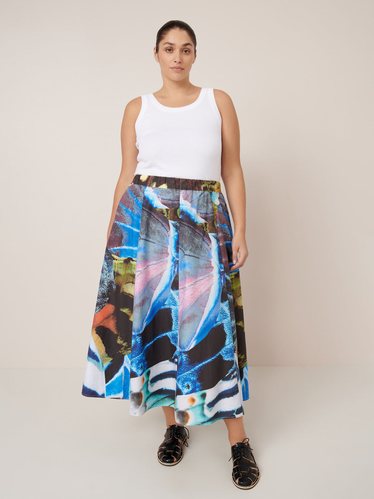 A woman stands against a plain background wearing a white tank top, the Kowtow Mosaic Skirt – Monarch with a high-waisted colorful abstract print in a relaxed fit, and black shoes.