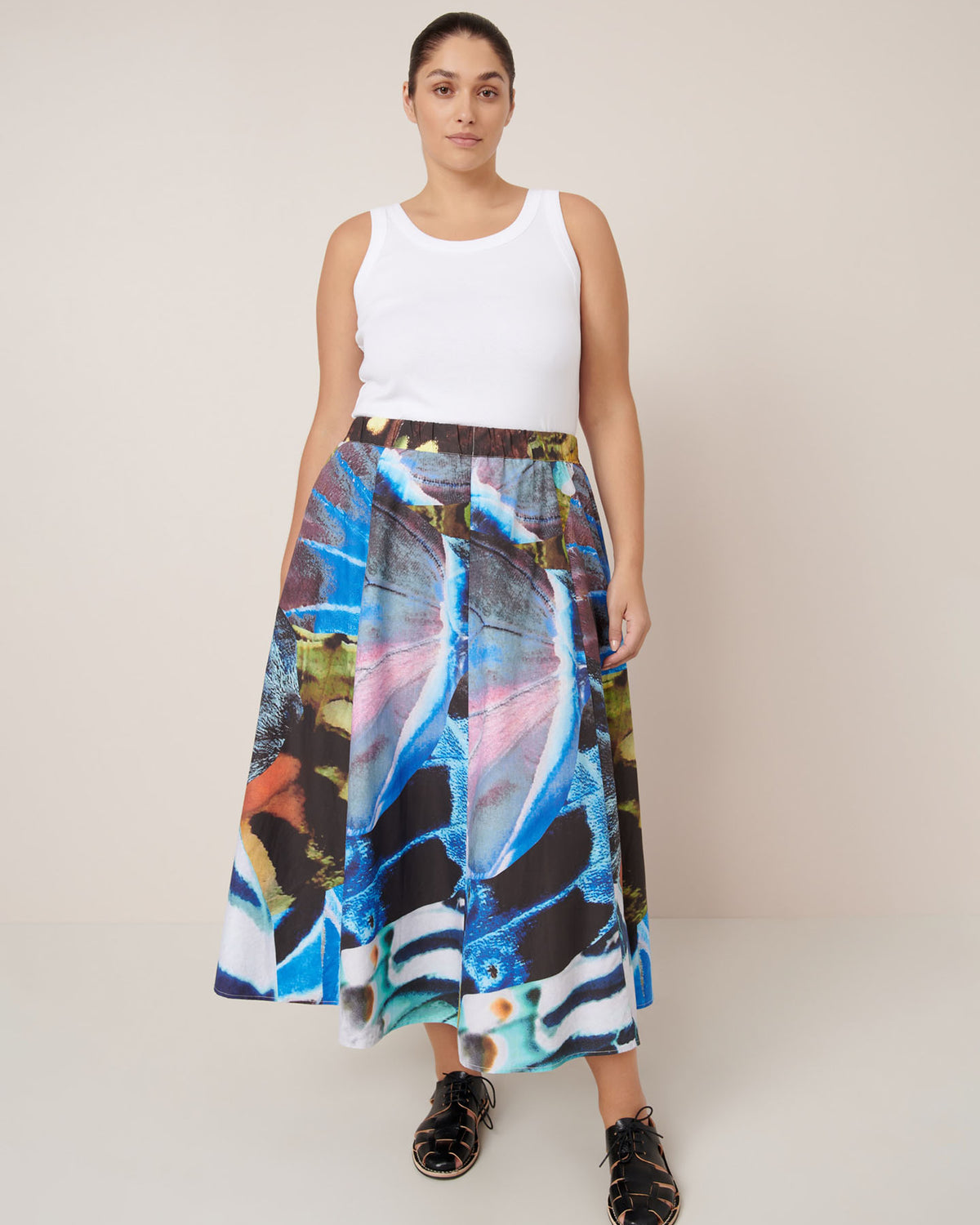 A woman stands against a plain background wearing a white tank top, the Kowtow Mosaic Skirt – Monarch with a high-waisted colorful abstract print in a relaxed fit, and black shoes.