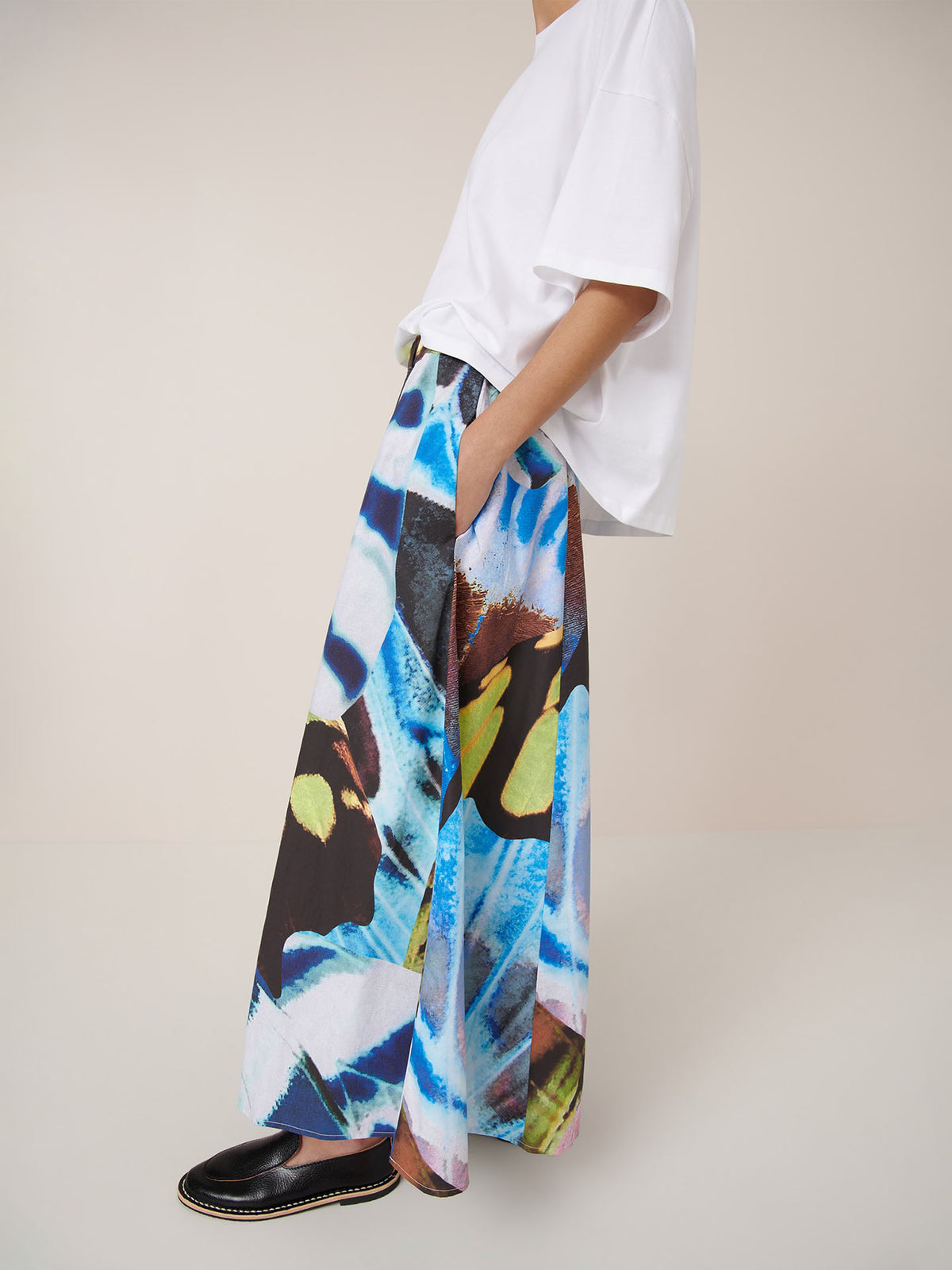 A person stands sideways wearing Kowtow’s Mosaic Skirt – Monarch, styled for a relaxed fit with a loose white t-shirt and black slip-on shoes.