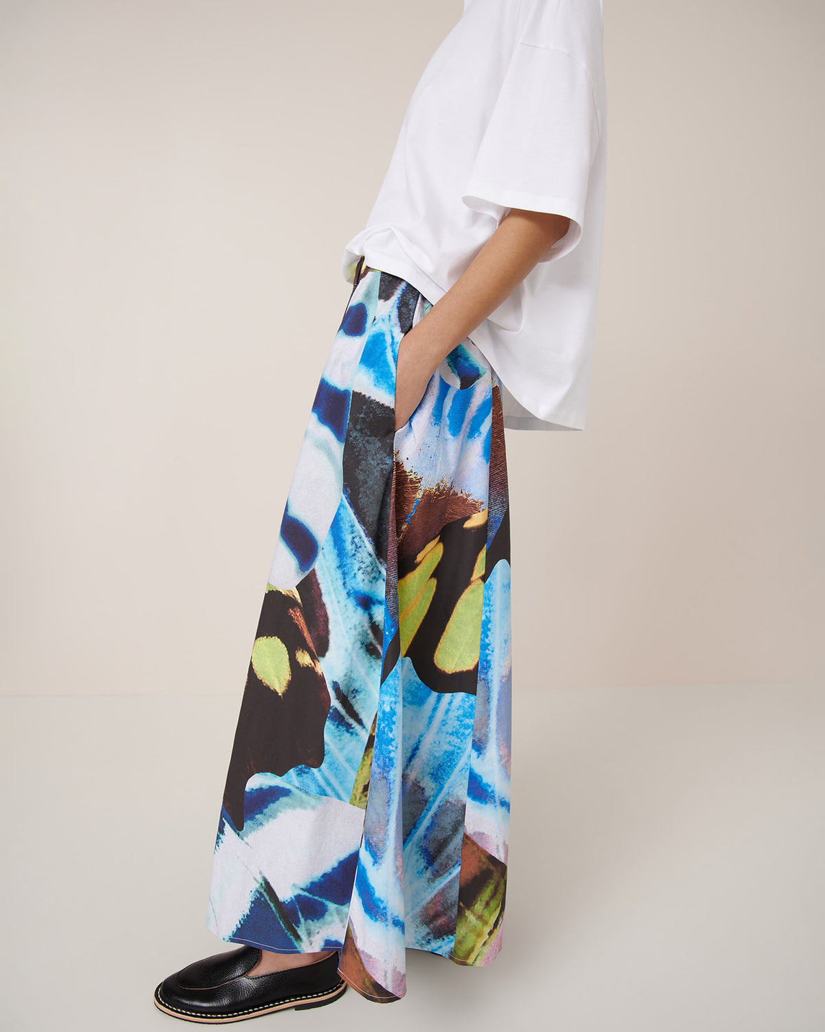 A person stands sideways wearing Kowtow’s Mosaic Skirt – Monarch, styled for a relaxed fit with a loose white t-shirt and black slip-on shoes.