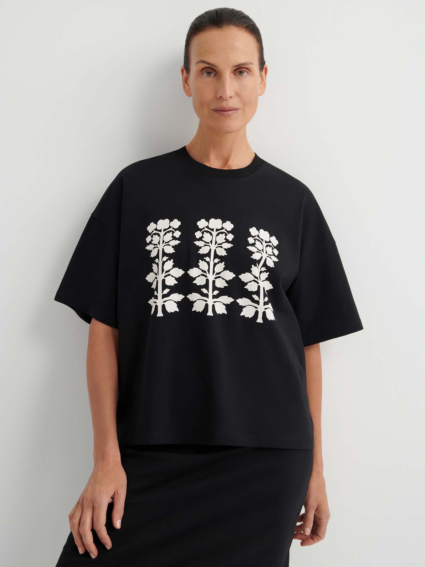 A woman wearing the Motif Tee – Black, an oversized boxy top with drop shoulder sleeves and a white floral print, stands against a plain white background.