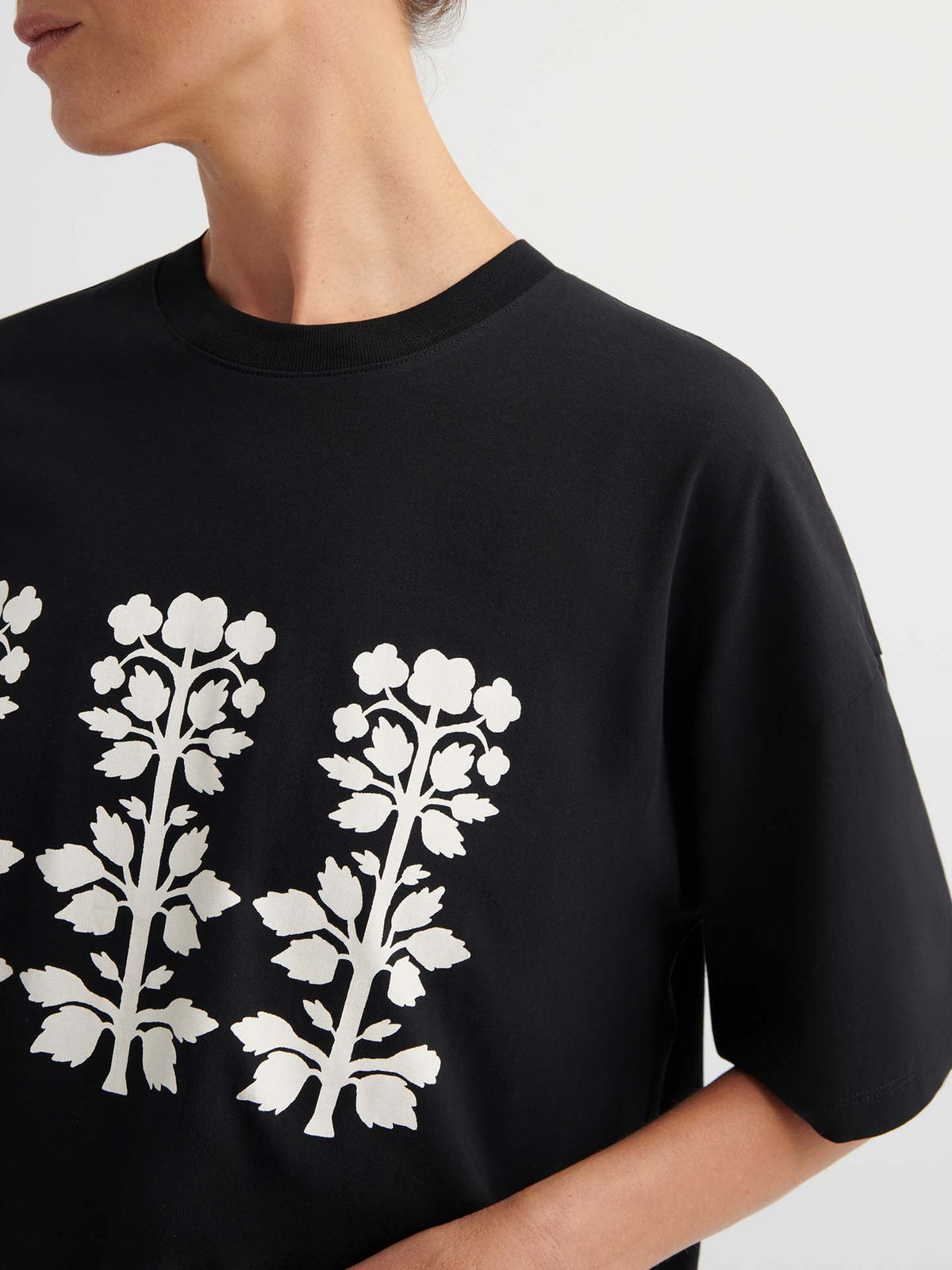 A person wears the Motif Tee – Black, featuring a white floral print, oversized boxy fit, and drop shoulder sleeves, standing against a plain background.