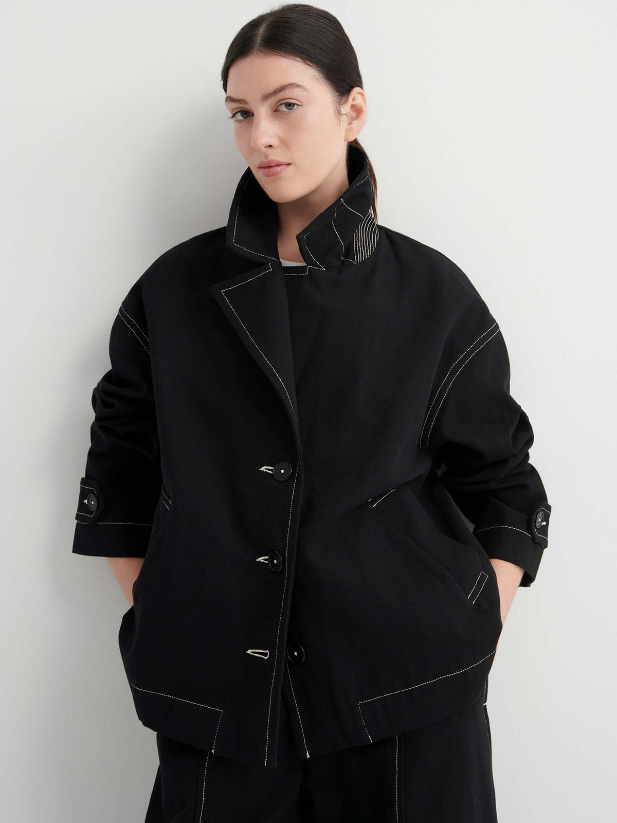 A woman stands against a plain light background, wearing the Mural Jacket – Black featuring contrasting white stitching and drop shoulder sleeves.