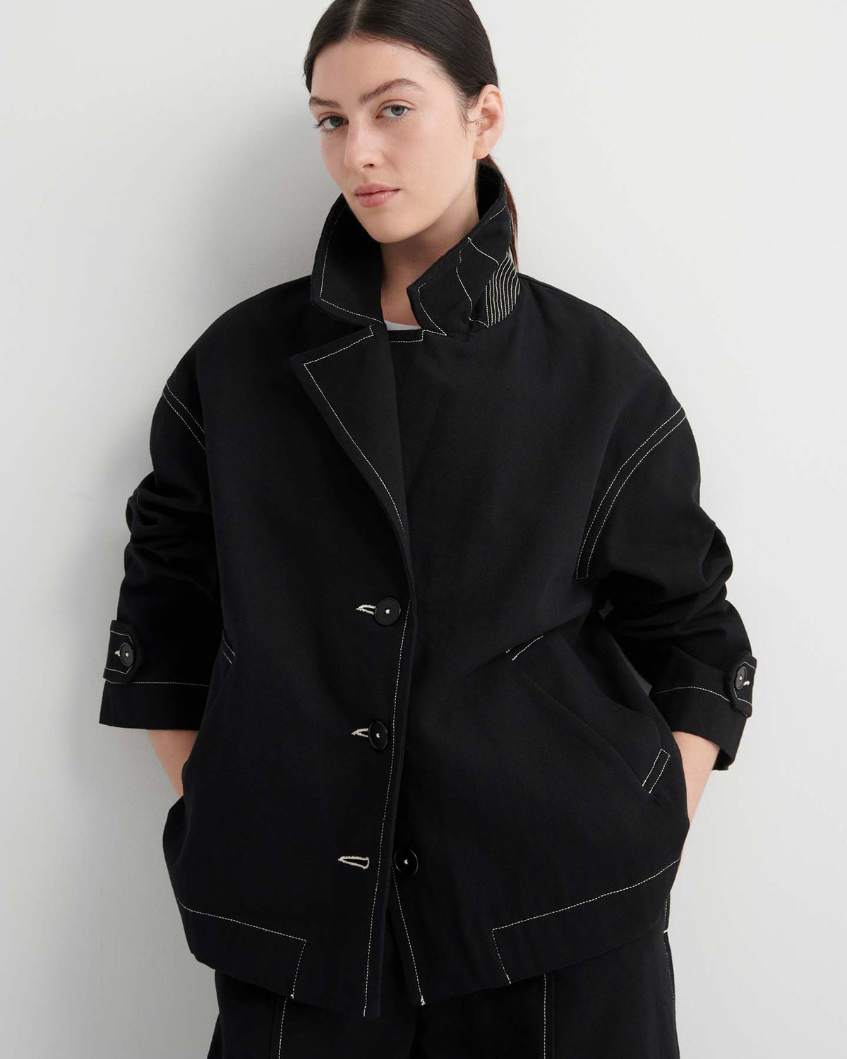 A woman stands against a plain light background, wearing the Mural Jacket – Black featuring contrasting white stitching and drop shoulder sleeves.
