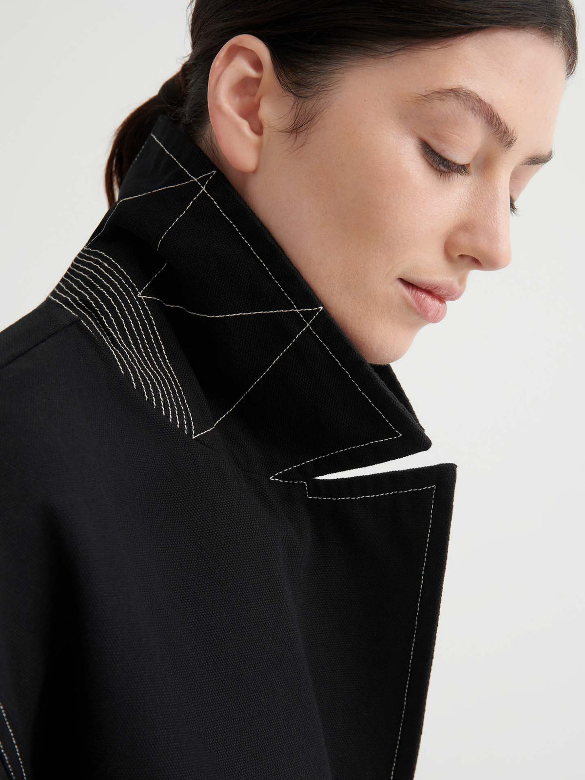 Someone with dark hair wears the Mural Jacket – Black, an oversized piece with bold white stitching, shown from the side against a light background.