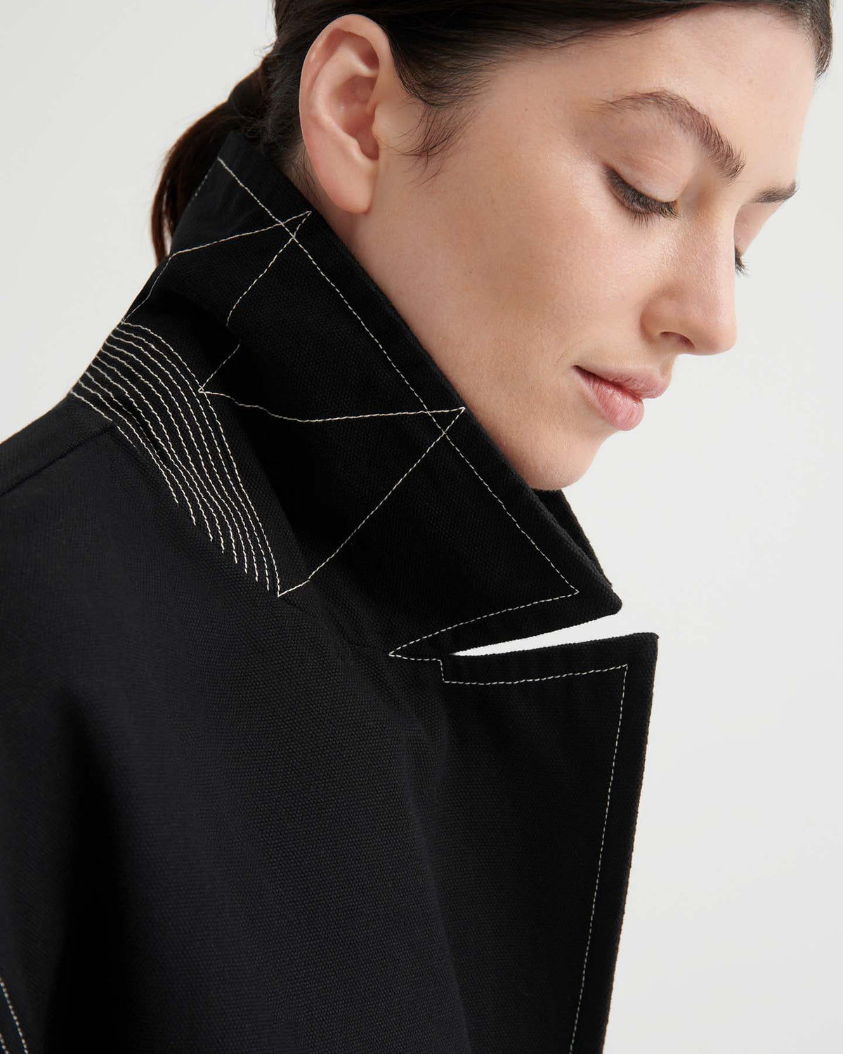 Someone with dark hair wears the Mural Jacket – Black, an oversized piece with bold white stitching, shown from the side against a light background.