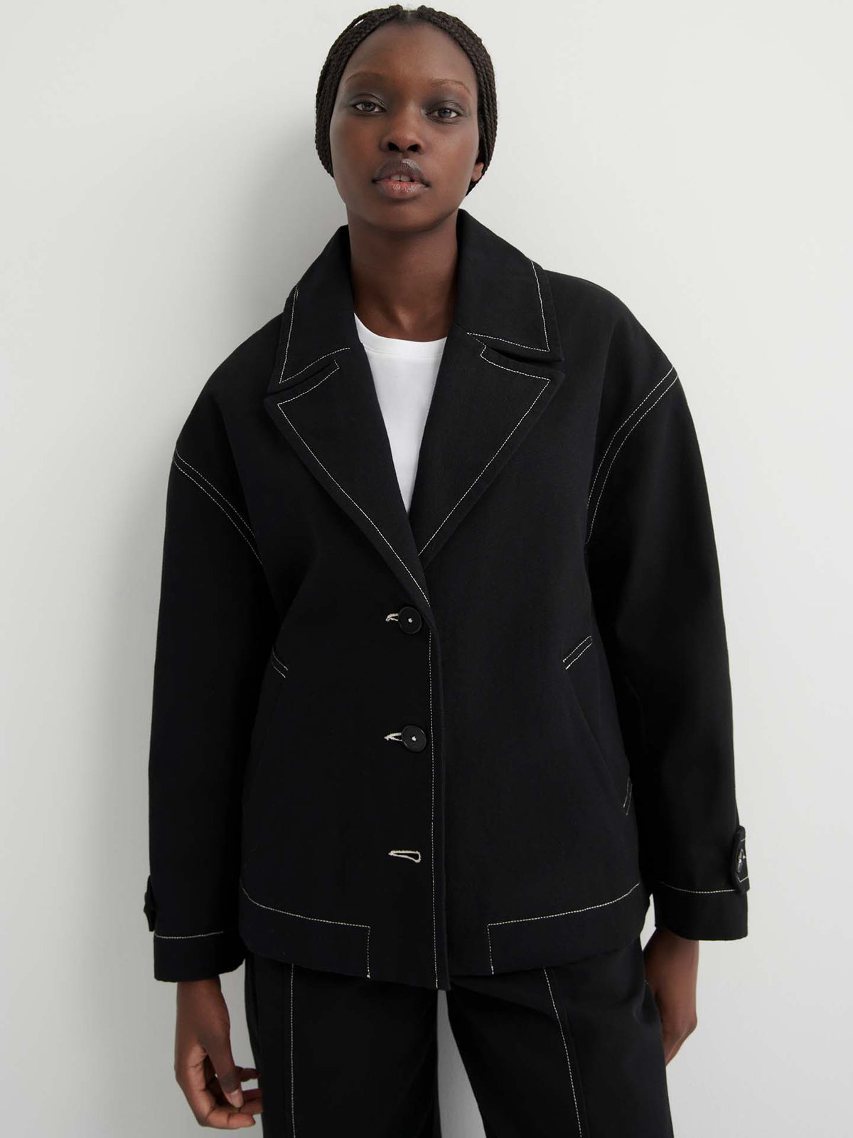 Someone stands against a plain light background wearing the oversized Mural Jacket – Black, featuring drop shoulder sleeves and white stitching, layered over a white shirt.
