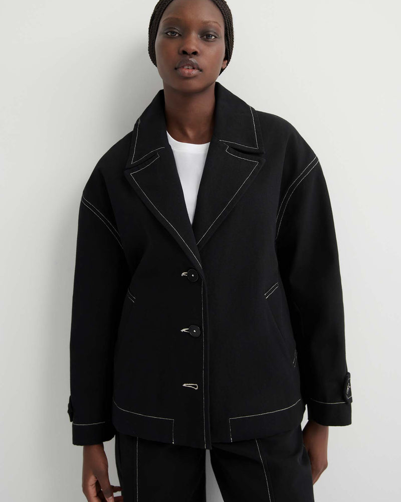 Someone stands against a plain light background wearing the oversized Mural Jacket – Black, featuring drop shoulder sleeves and white stitching, layered over a white shirt.