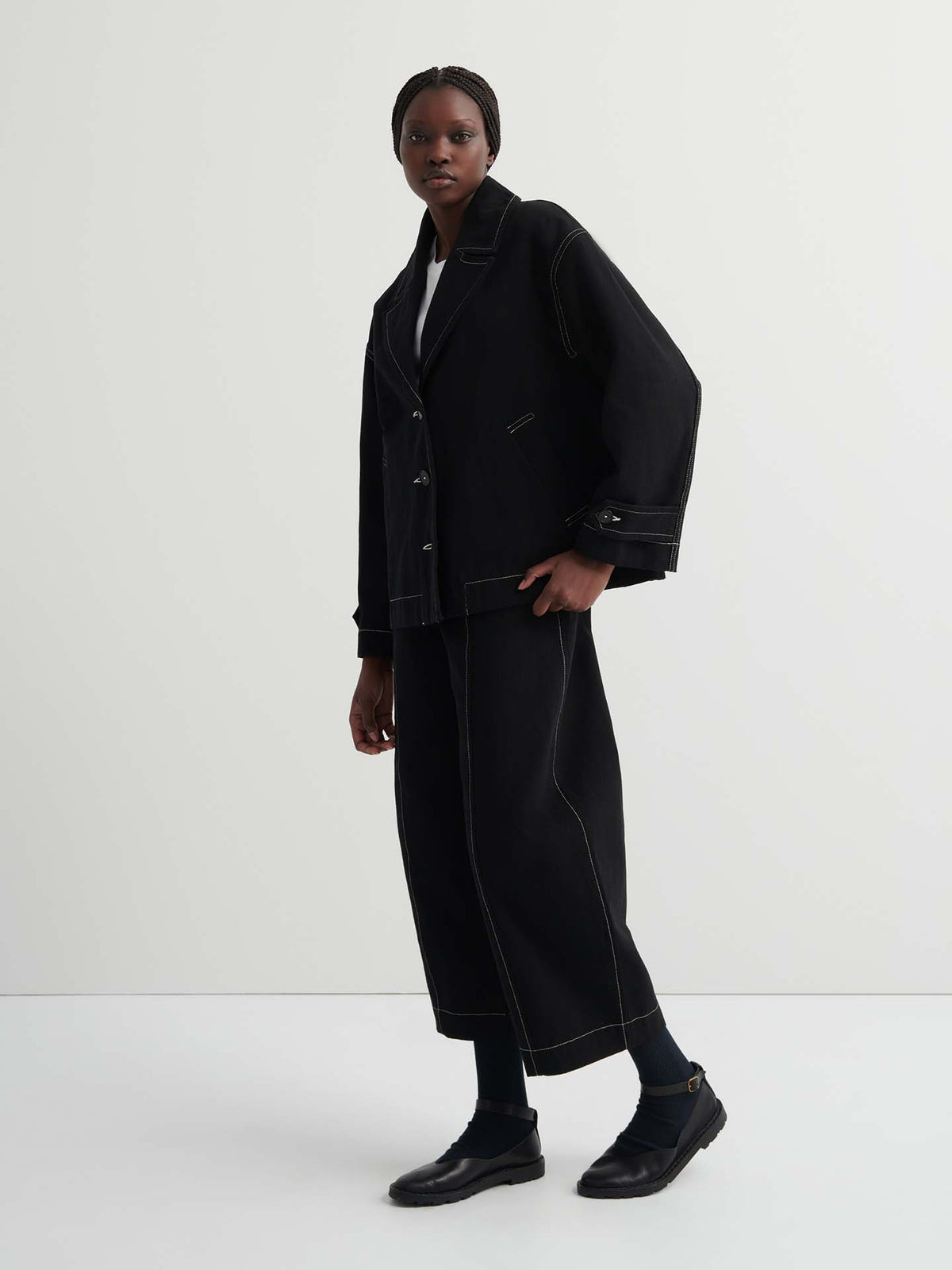 A person in the black Mural Jacket with a boxy, oversized fit, paired with matching wide-leg pants with contrast stitching, black shoes, and braided hair stands against a plain white background.